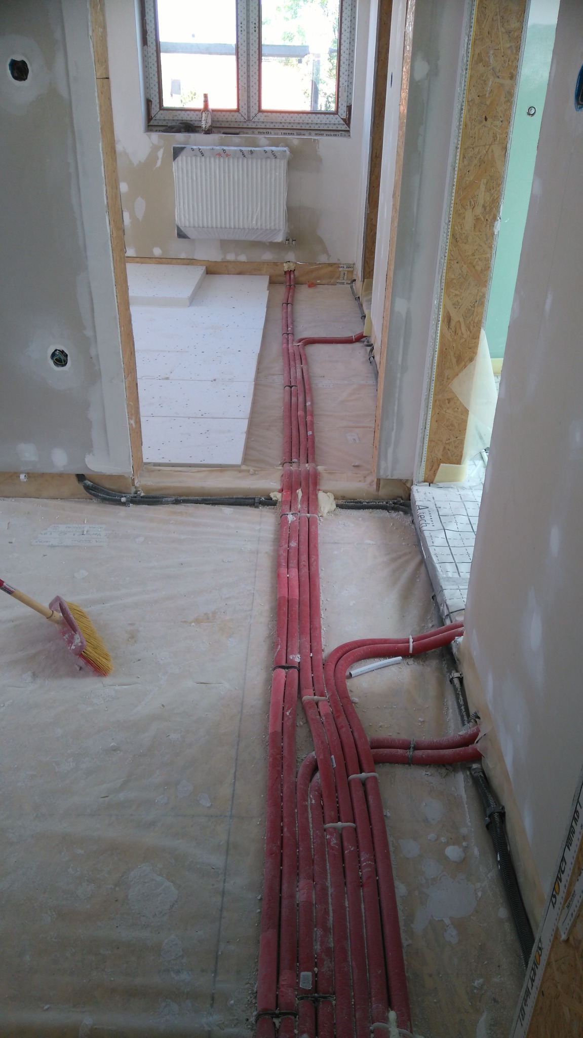 Montaż ogrzewania podłogowego: czerwone rury grzewcze ułożone na izolacji podłogowej w surowym wnętrzu, widoczny fragment ściany z płyt karton-gips i przygotowane przejście do kolejnego...