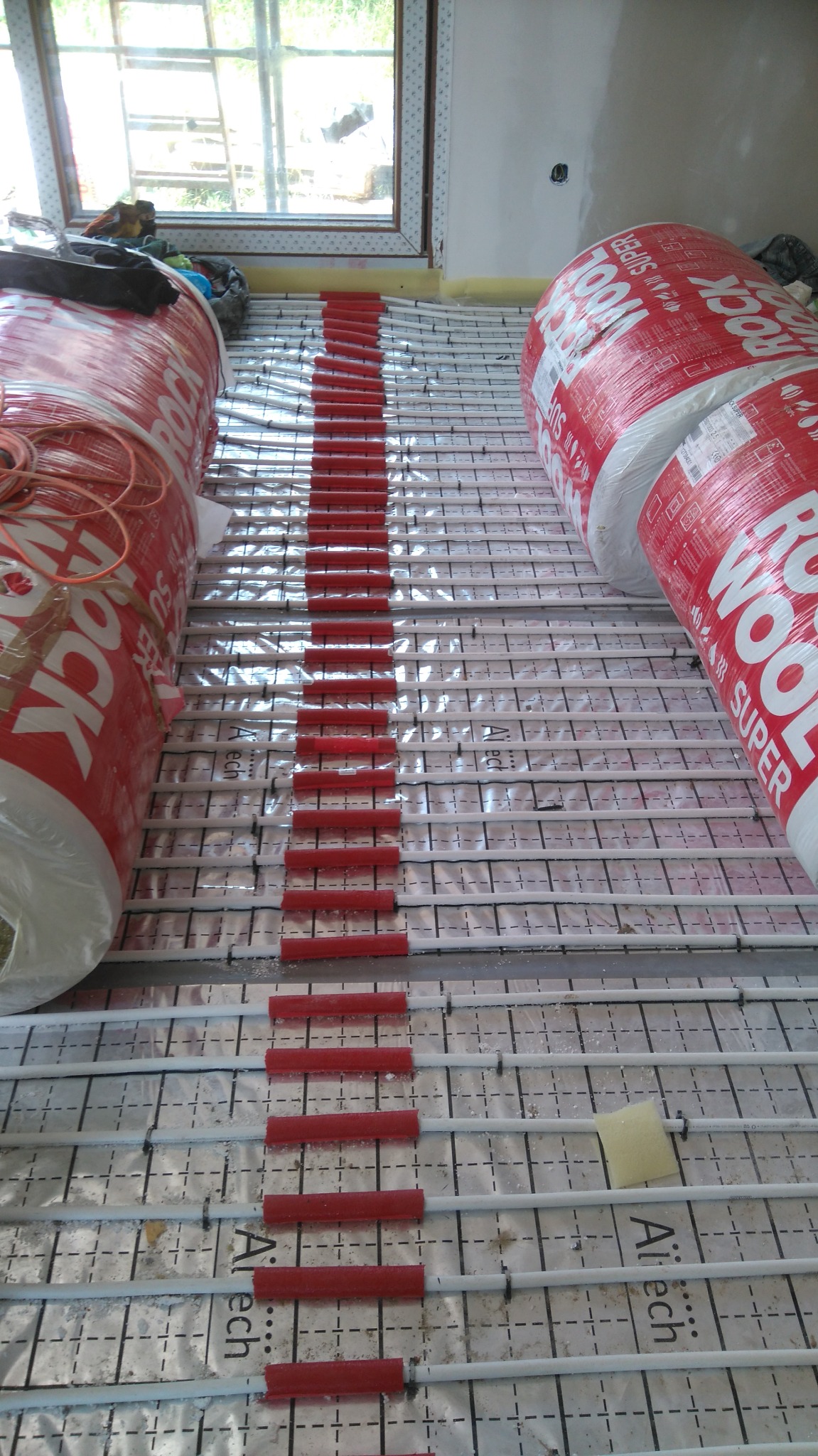 Instalacja ogrzewania podłogowego: białe rurki grzewcze zamocowane na folii izolacyjnej z czerwonymi elementami mocującymi, obok leżą zrolowane materiały izolacyjne z widocznym logo producenta.