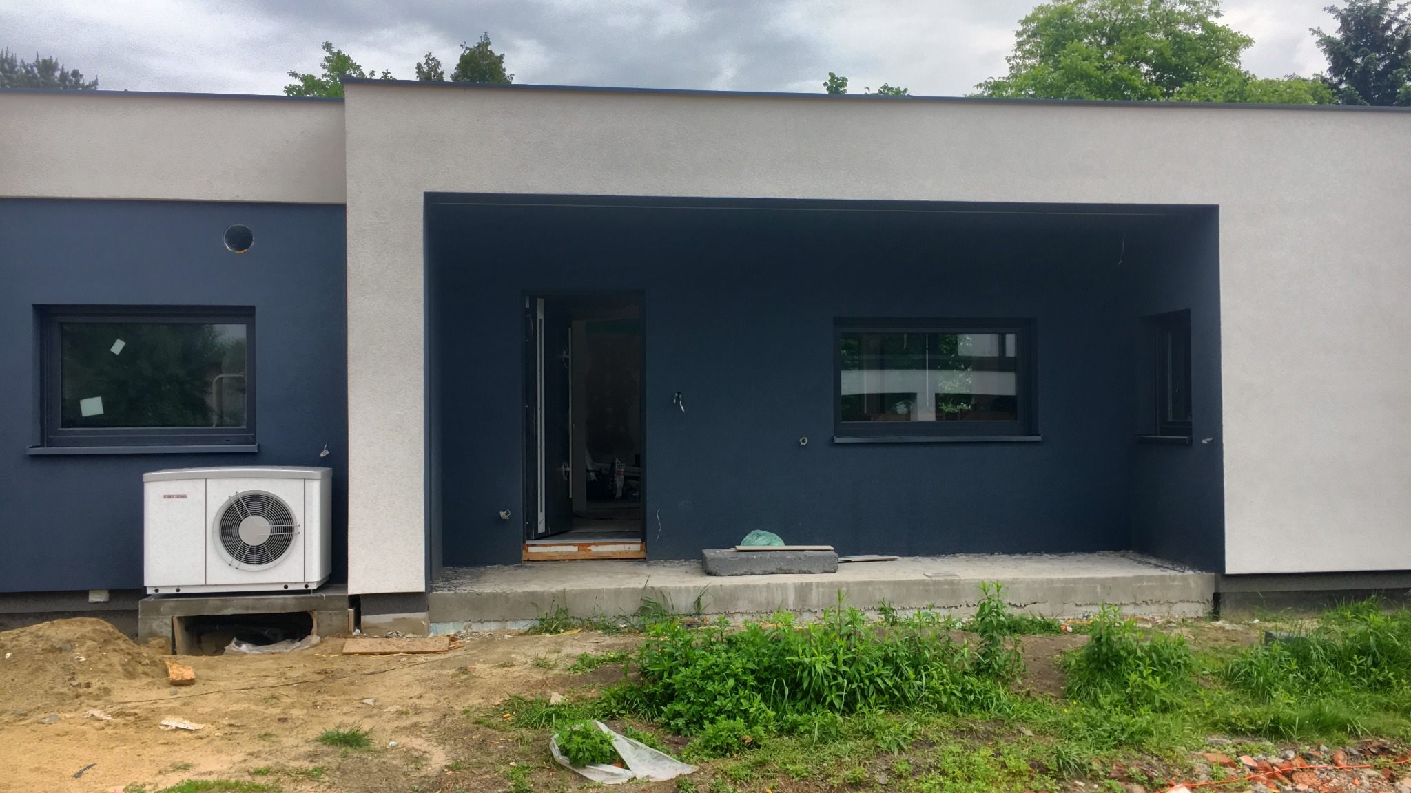 Nowoczesny dom jednorodzinny w trakcie wykończenia, z zamontowaną jednostką zewnętrzną pompy ciepła, widoczne niedokończone prace ziemne wokół budynku.