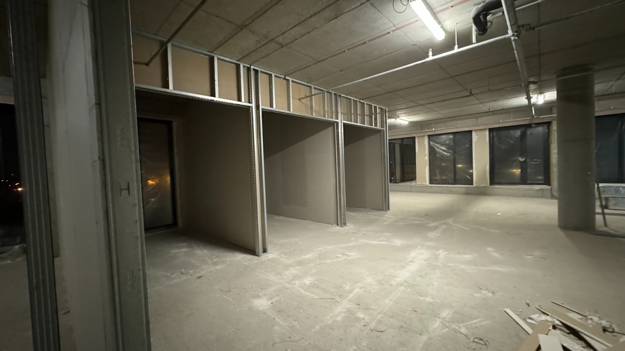 Wnętrze w trakcie prac wykończeniowych, widoczne metalowe profile konstrukcyjne i płyty gipsowo-kartonowe tworzące ścianki działowe w dużym pomieszczeniu z betonowym stropem i oknami.