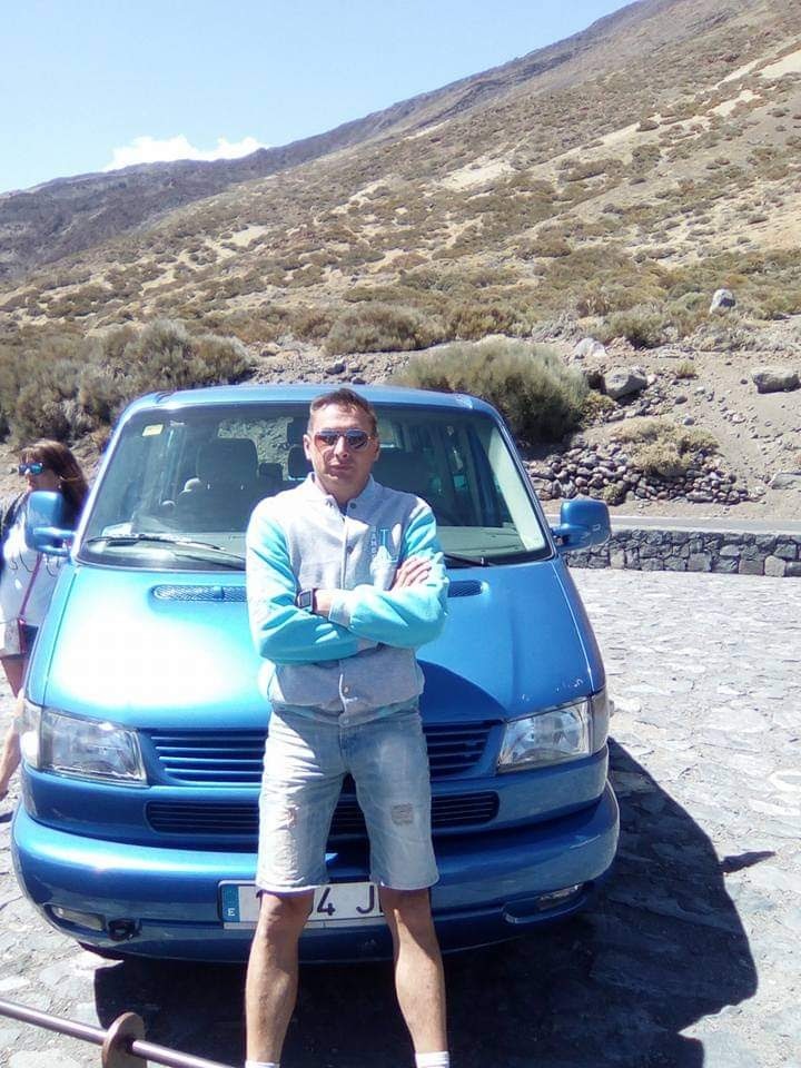 Mężczyzna w okularach przeciwsłonecznych stoi przed niebieskim busem z widokiem na górzysty krajobraz w tle.