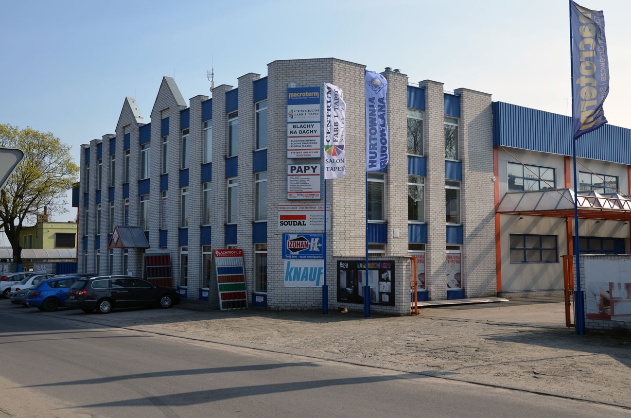 Budynek składu budowlanego z ekspozycją blach trapezowych i reklamami producentów materiałów budowlanych, widoczne flagi reklamowe.