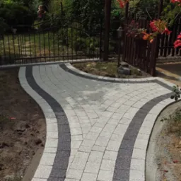 Ścieżka z jasnoszarej kostki brukowej z ciemnoszarymi pasami, rozwidlająca się w ogrodzie z altaną w tle.