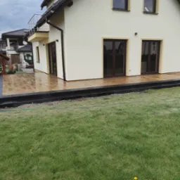 Nowoczesny taras z jasnobrązowej kostki brukowej przed domem z beżową elewacją, obrzeża z ciemnoszarej kostki, widoczny fragment trawnika i altana w tle.