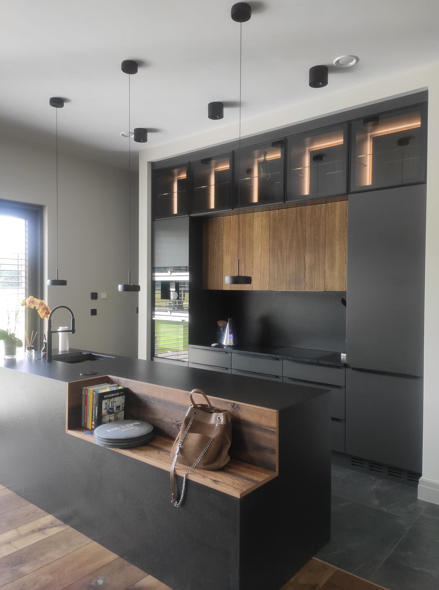 Nowoczesna kuchnia z wyspą, czarne matowe fronty szafek, drewniane akcenty na ścianie i w wyspie, oświetlenie punktowe, torba i książki na półce w wyspie.