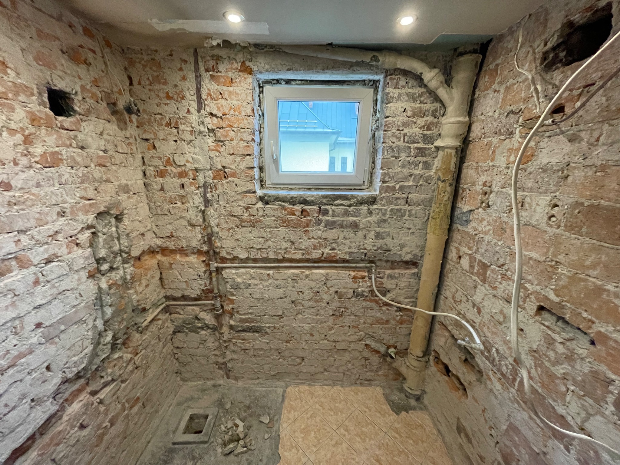 Surowy stan łazienki w trakcie remontu: odsłonięta cegła, instalacje, fragmenty płytek podłogowych i okno z widokiem na budynek.