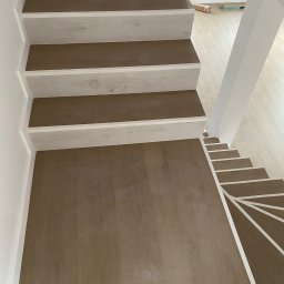 Saranda - montaż podłóg Rafał Trębski - Widok z góry na jasne schody drewniane z brązowymi stopniami i białymi podstopnicami, z widocznym fragmentem podłogi i kartonami w tle.
