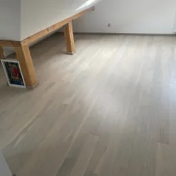 Jasna, drewniana podłoga w pokoju na poddaszu z drewnianymi belkami stropowymi. Widoczny fragment instalacji w ścianie i grzejnik.