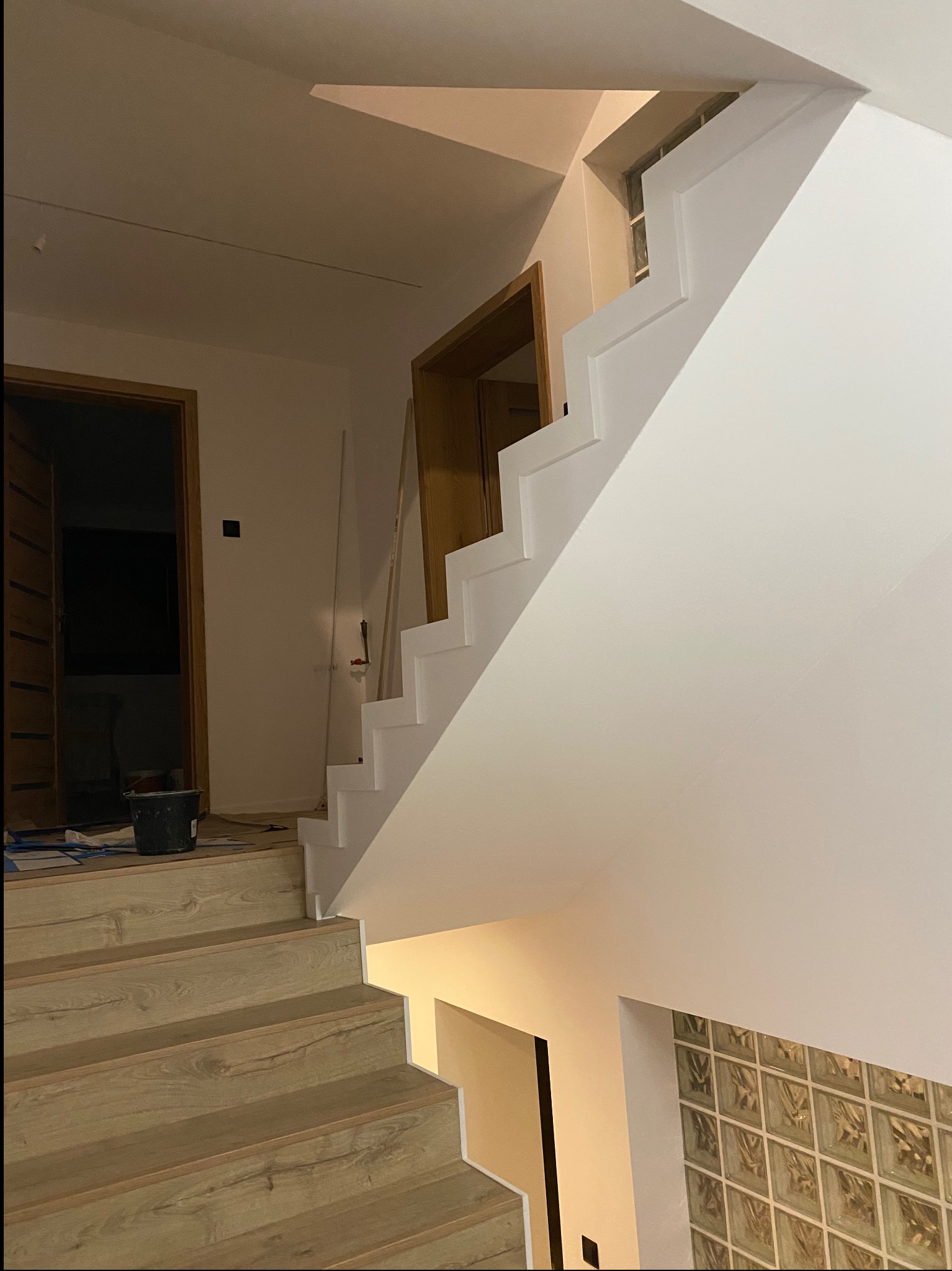 Wnętrze domu w trakcie remontu: drewniane stopnie schodów prowadzące do góry, pomalowane na biało ściany i balustrada, widoczny kubeł i narzędzia malarskie.