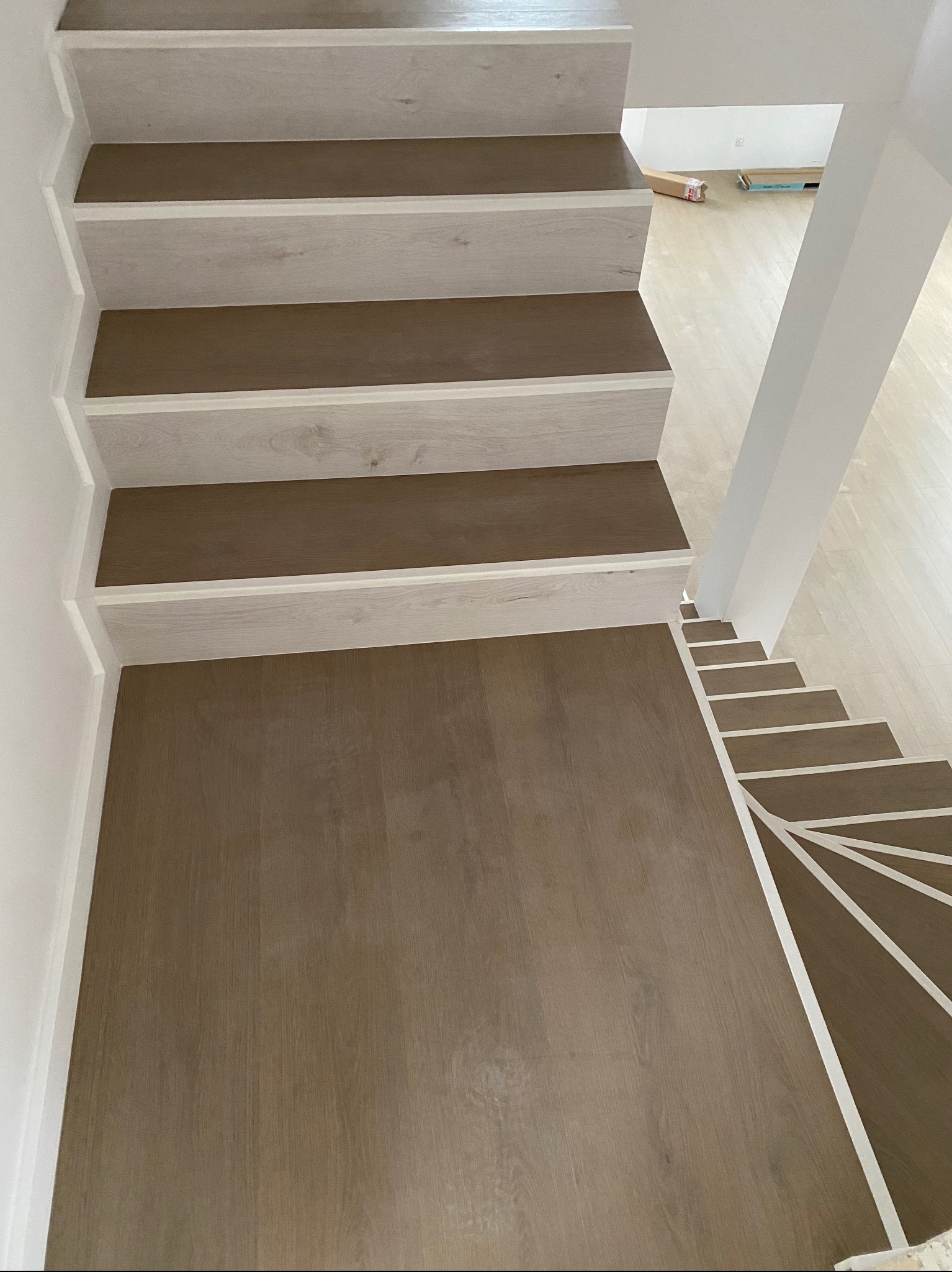 Widok z góry na jasne schody drewniane z brązowymi stopniami i białymi podstopnicami, z widocznym fragmentem podłogi i kartonami w tle.