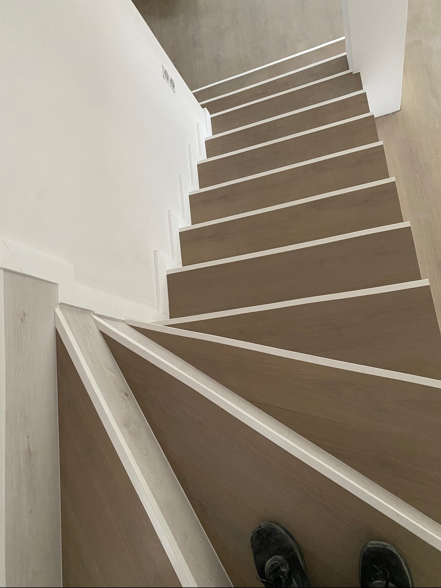Widok z góry na spiralne schody z drewnianymi stopniami i białymi listwami wykończeniowymi; na dolnym stopniu widoczne buty osoby fotografującej.