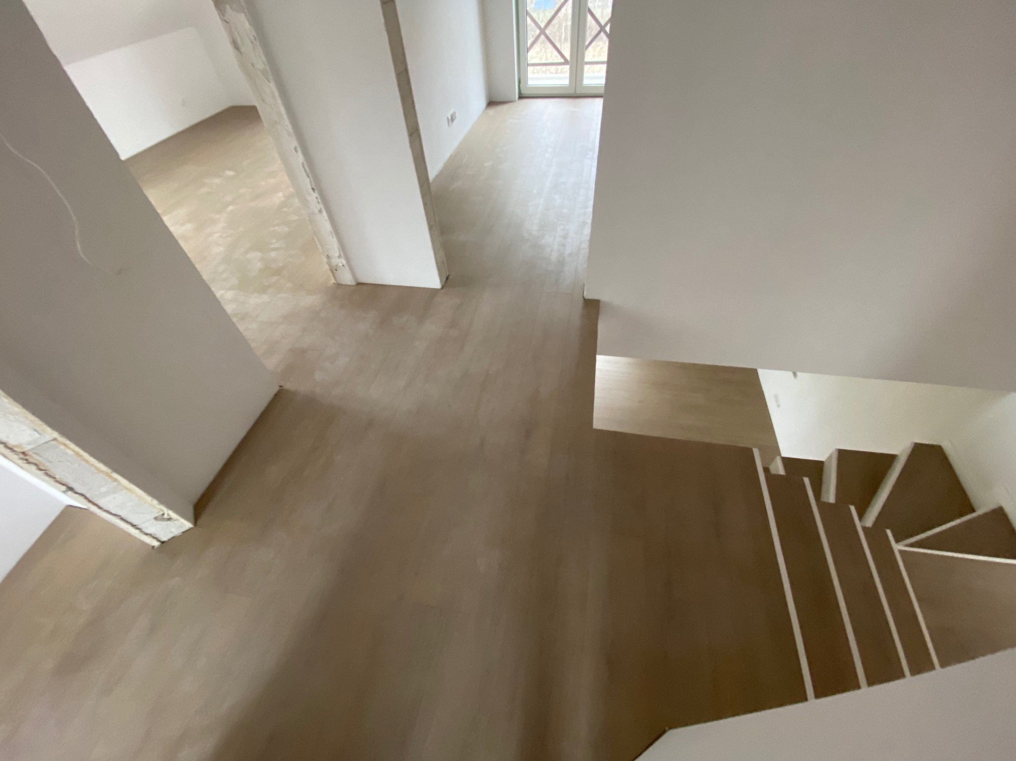 Widok z góry na nowo wykończone wnętrze w Skierniewicach z jasnymi panelami podłogowymi, schody z drewnianymi stopniami prowadzące w dół, białe ściany i widoczne okno w oddali.