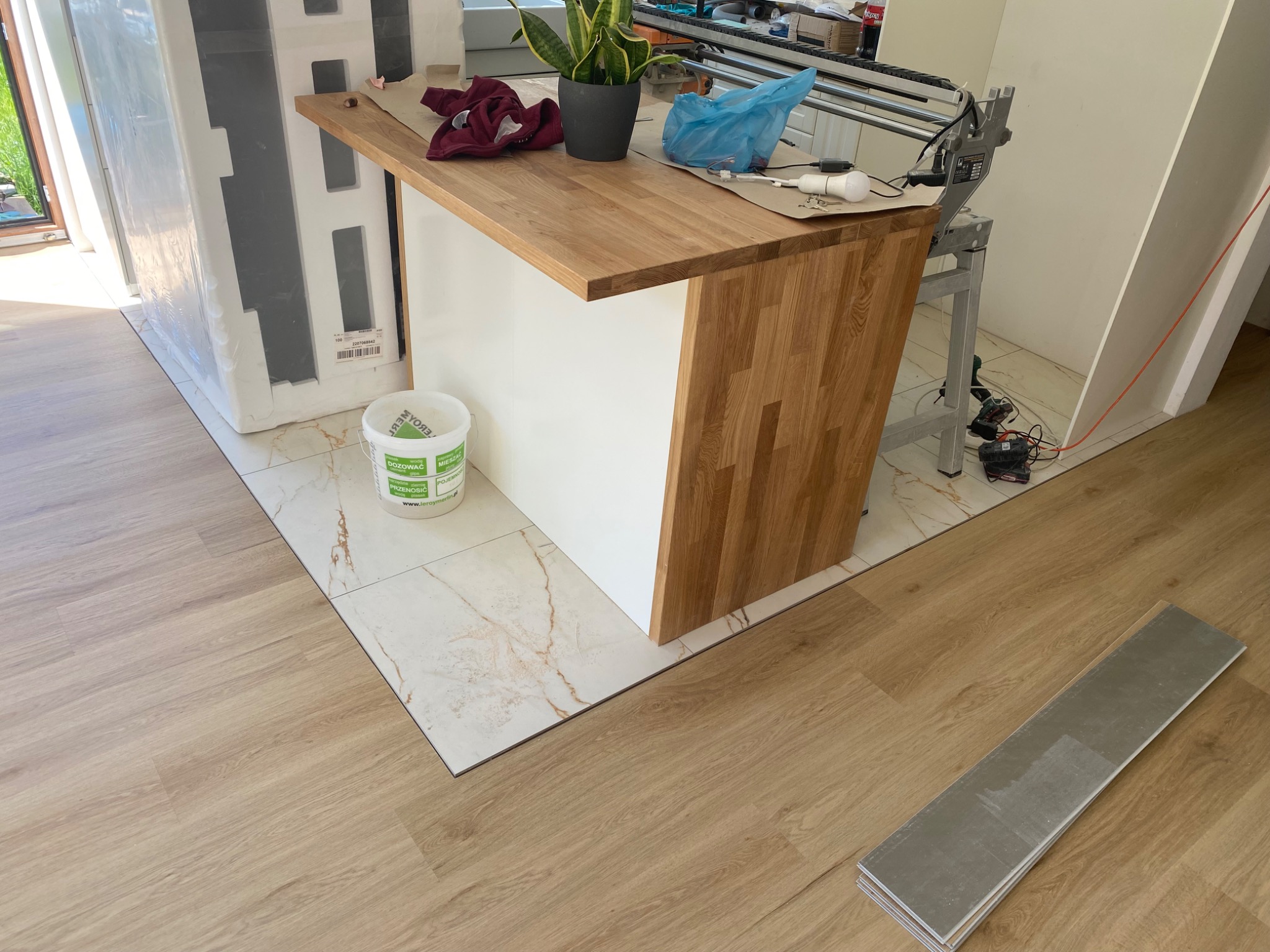 Fragment pomieszczenia z ułożonymi panelami podłogowymi w odcieniach drewna, widoczny fragment wyspy kuchennej i narzędzia do montażu paneli.