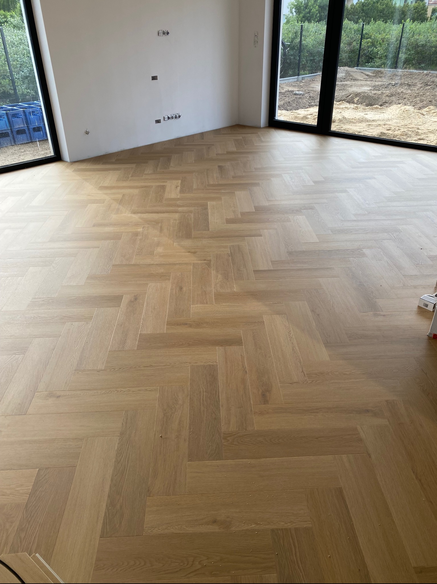 Jasny parkiet ułożony we wzór jodełki w nowym wnętrzu z dużymi oknami. Widoczne gniazdka elektryczne na ścianie i fragment skrzynki na zewnątrz okna.