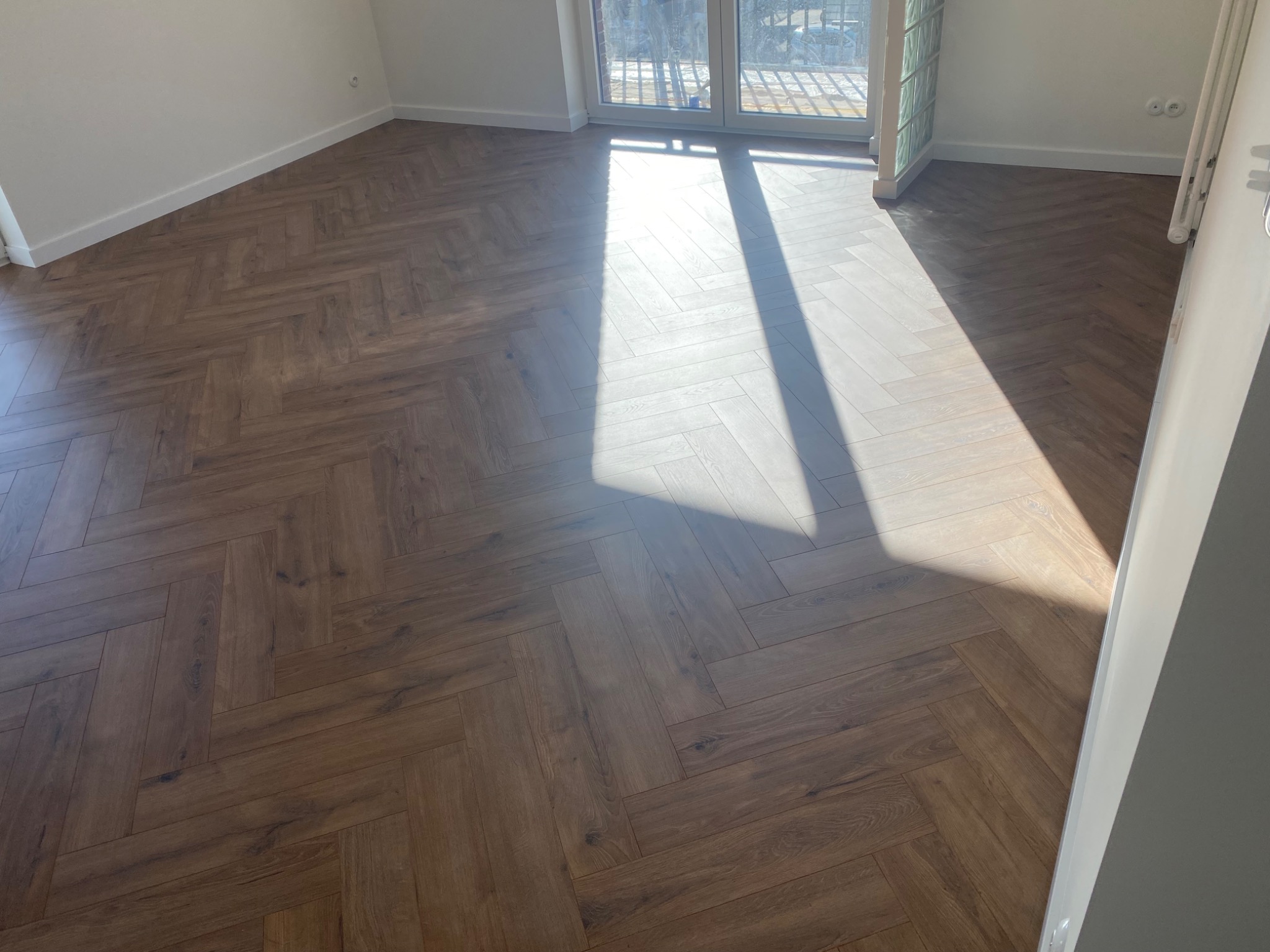 Ułożona jodełka z paneli w jasnym pokoju, z widocznym fragmentem okna i padającym światłem słonecznym, podkreślającym wzór podłogi.