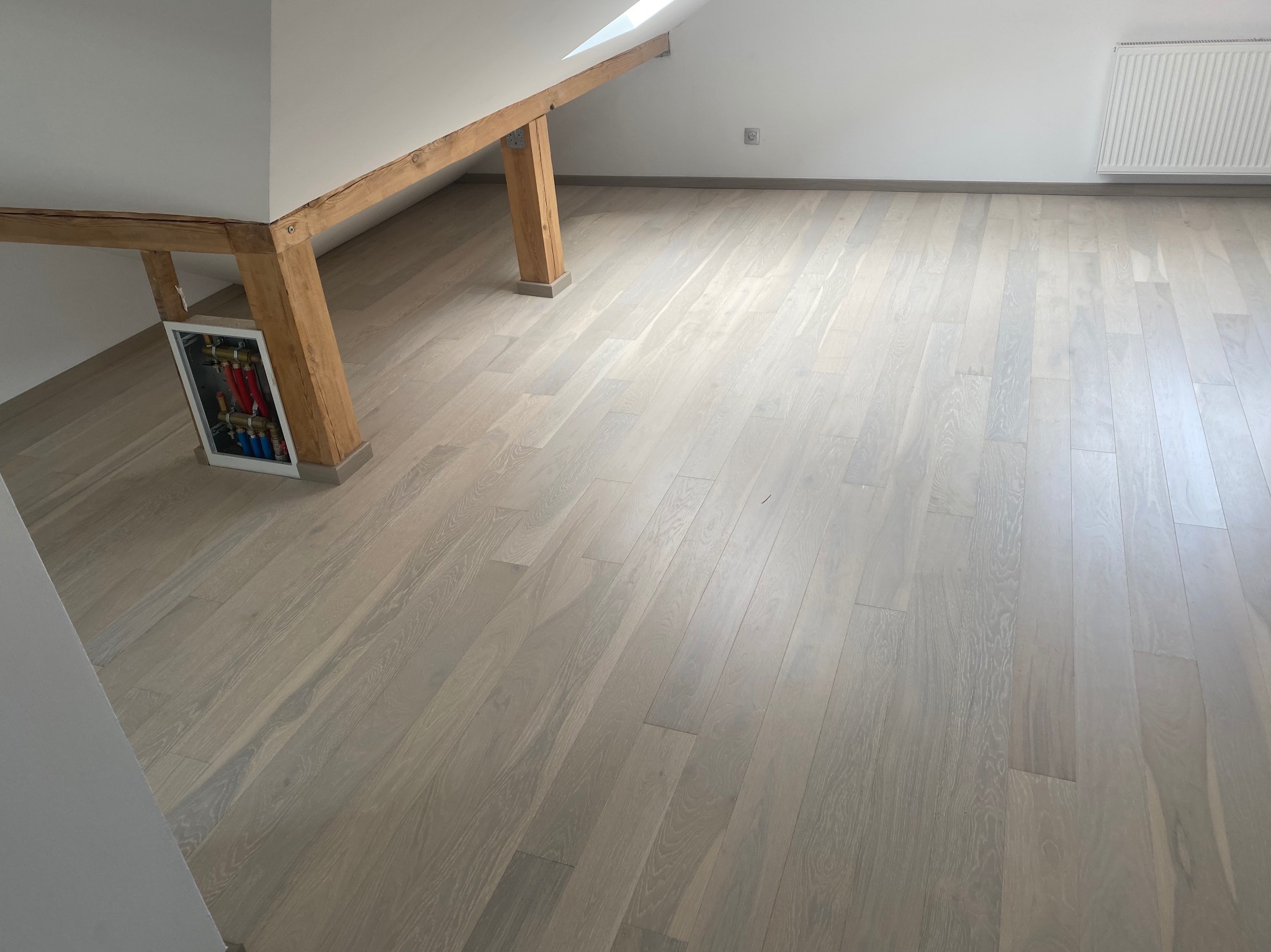 Jasna, drewniana podłoga w pokoju na poddaszu z drewnianymi belkami stropowymi. Widoczny fragment instalacji w ścianie i grzejnik.