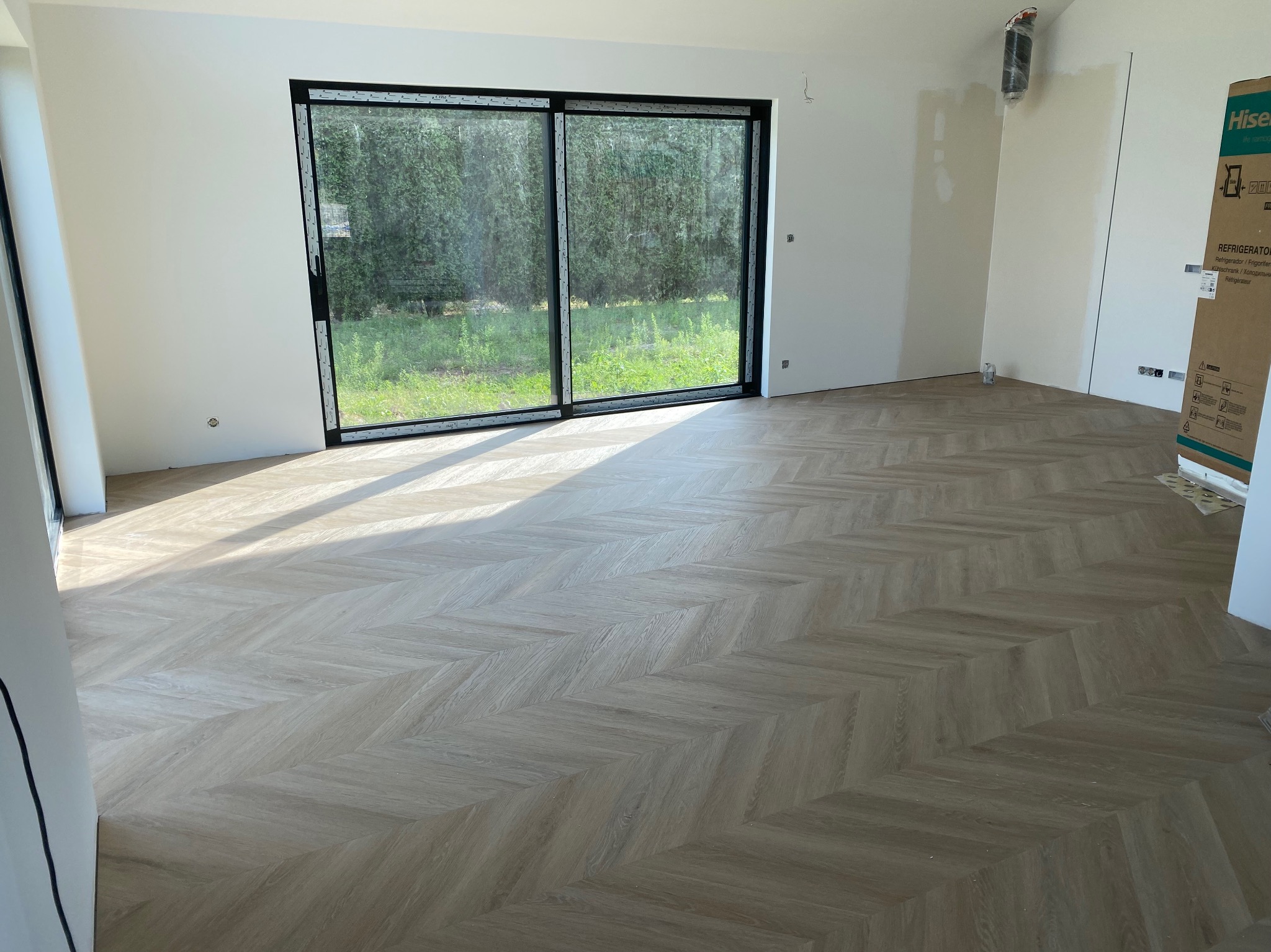 Jasna podłoga ułożona we wzór jodełki w nowym wnętrzu z dużym oknem przesuwnym i światłem słonecznym wpadającym do środka. Widoczny karton po lodówce.