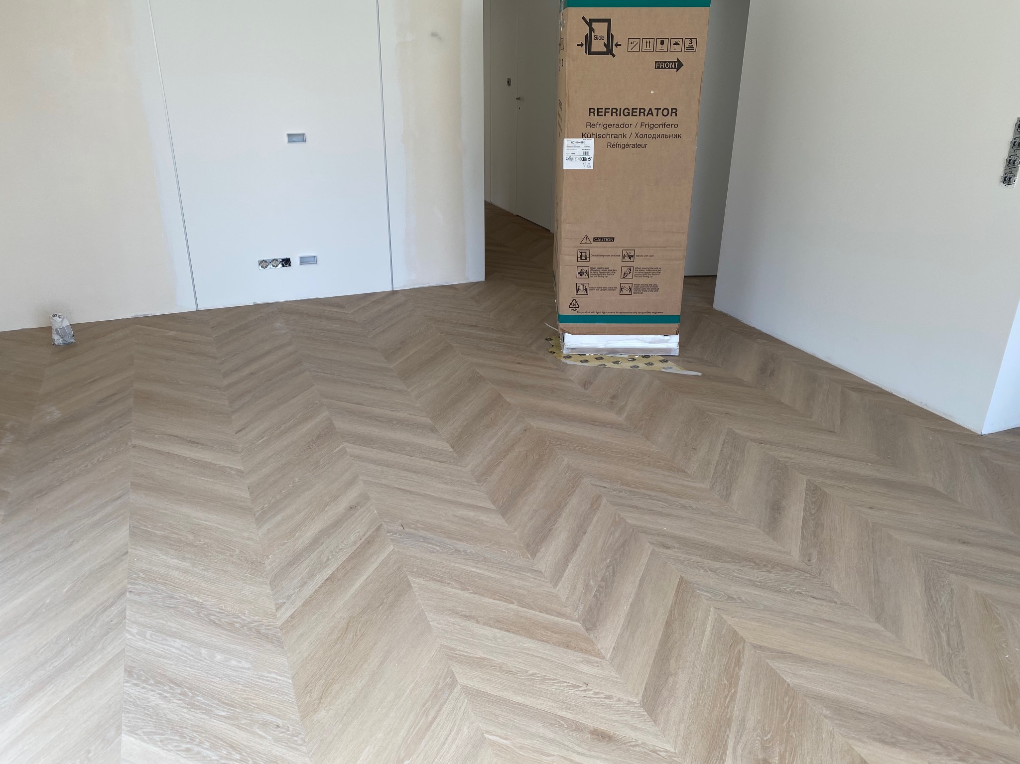 Ułożony parkiet w jodełkę francuską w jasnym odcieniu drewna, z kartonem po lodówce w tle. Widoczne gniazdka elektryczne na ścianie i fragment otwartego przejścia.