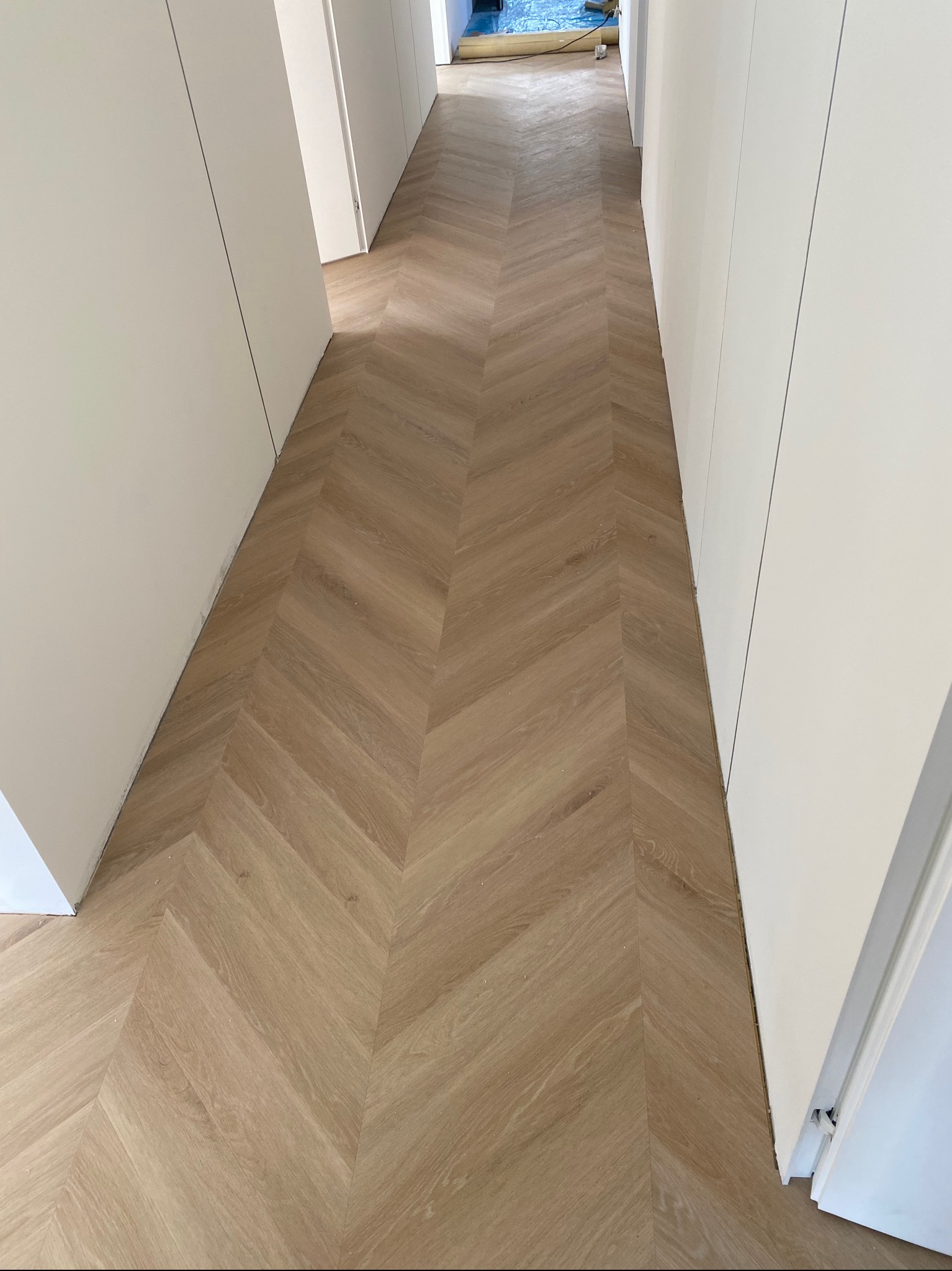 Jasne panele ułożone we wzór jodełki francuskiej w korytarzu z białymi ścianami i drzwiami. Widok z góry, naturalne światło.