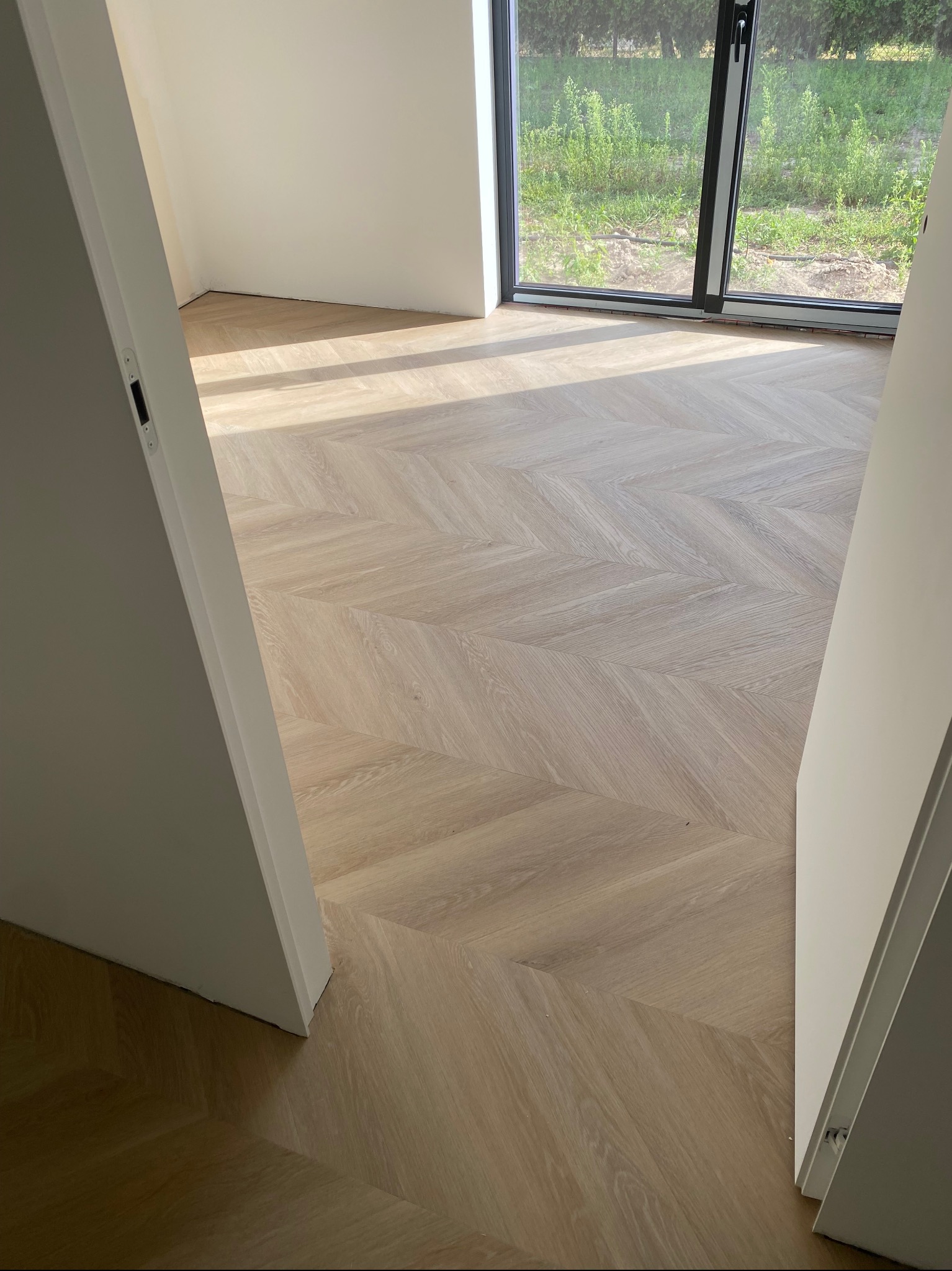 Jasna podłoga z paneli ułożonych we wzór jodełki francuskiej, widok z perspektywy drzwi. Duże okno z widokiem na zieleń w tle.