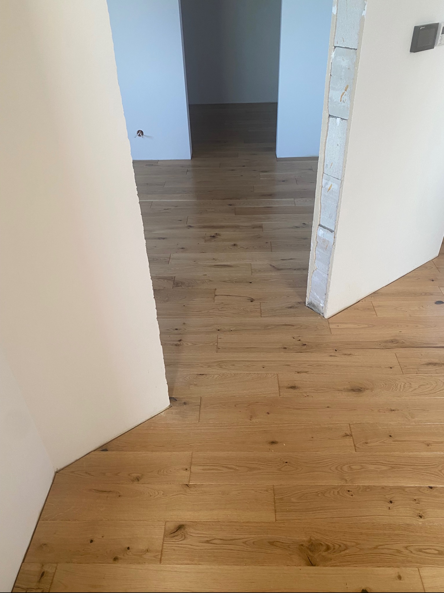 Jasne, drewniane deski podłogowe ułożone wzdłuż korytarza, widoczne przez otwarte drzwi. Ściany w odcieniach bieli. Fragment surowego wykończenia ościeżnicy.