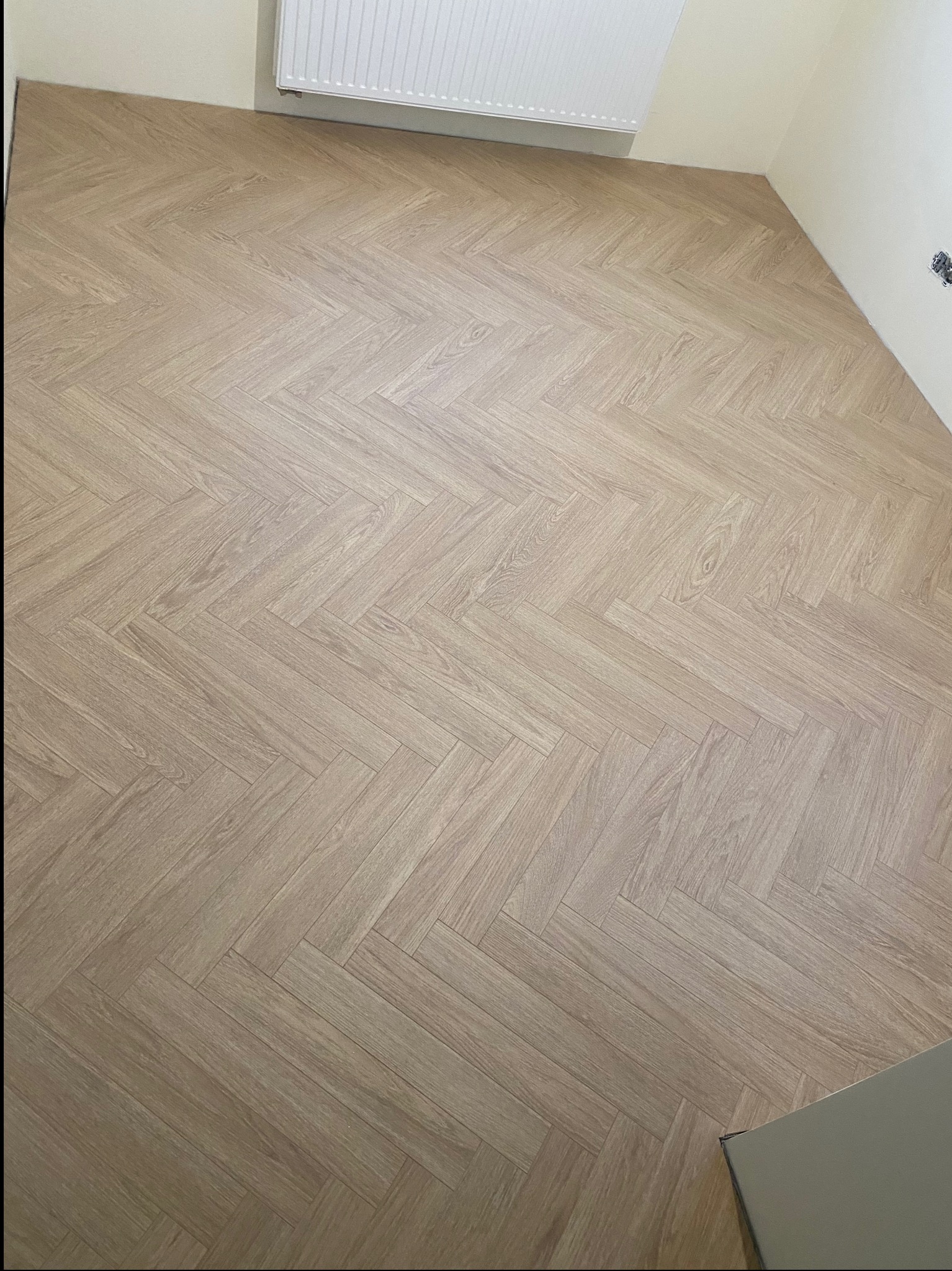 Jasne panele ułożone w jodełkę francuską, widok z góry na fragment pokoju z grzejnikiem i gniazdkiem elektrycznym. Nowoczesne wykończenie podłogi.