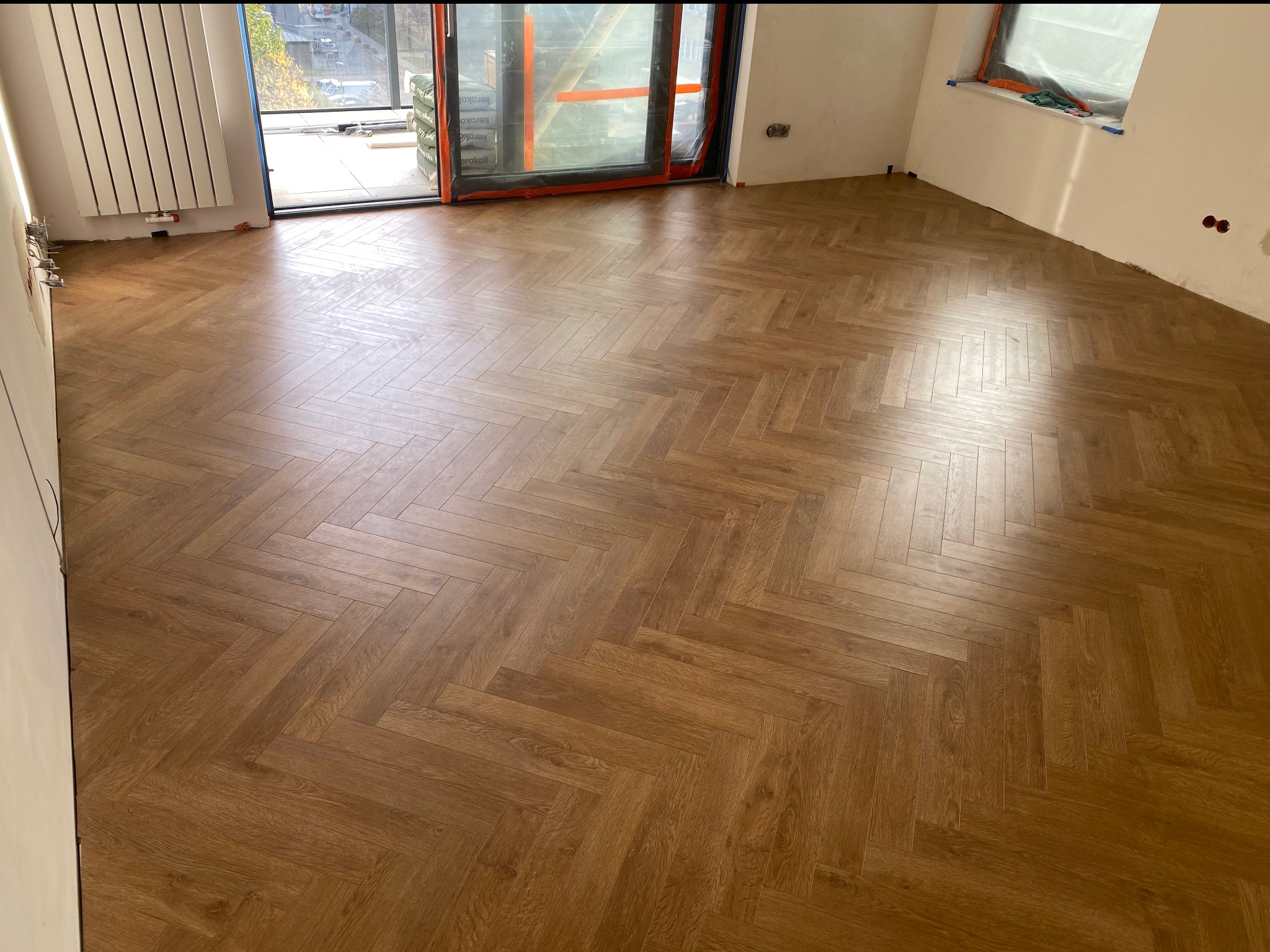 Ułożony parkiet w jodełkę francuską w jasnym pomieszczeniu z oknem balkonowym. Widoczne ślady po remoncie na ścianach. Podłoga z wyraźnym wzorem drewna.