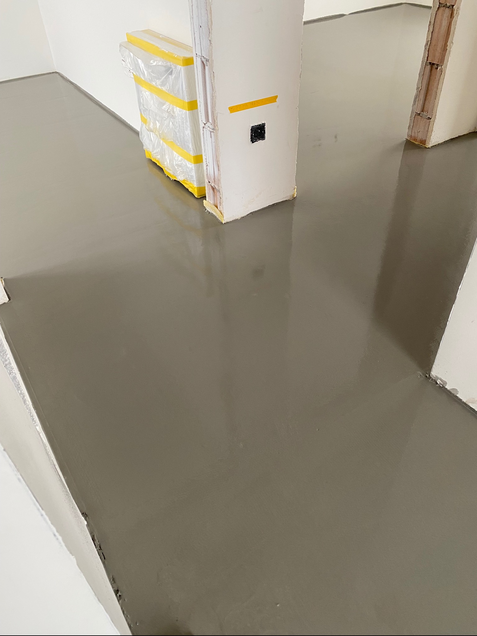 Świeżo wylana, gładka, szara posadzka żywiczna w pomieszczeniu w trakcie remontu. Widoczne zabezpieczone folią i taśmą elementy wyposażenia.