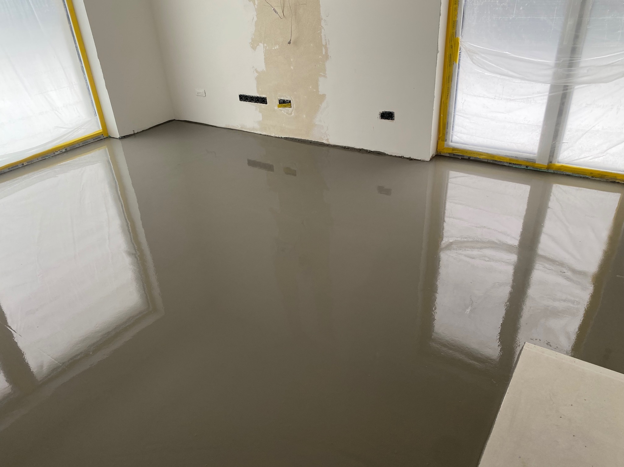 Wylana, mokra posadzka samopoziomująca w jasnym pomieszczeniu z oknami zabezpieczonymi folią malarską. Widoczne puszki elektryczne i przygotowane ściany.