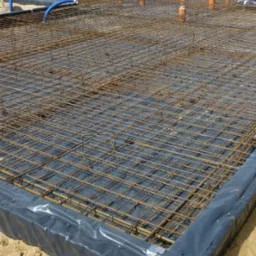 Przygotowanie zbrojenia pod płytę fundamentową: siatka z prętów stalowych na czarnej folii hydroizolacyjnej, widoczne pionowe rury kanalizacyjne i niebieski przewód doprowadzający wodę.