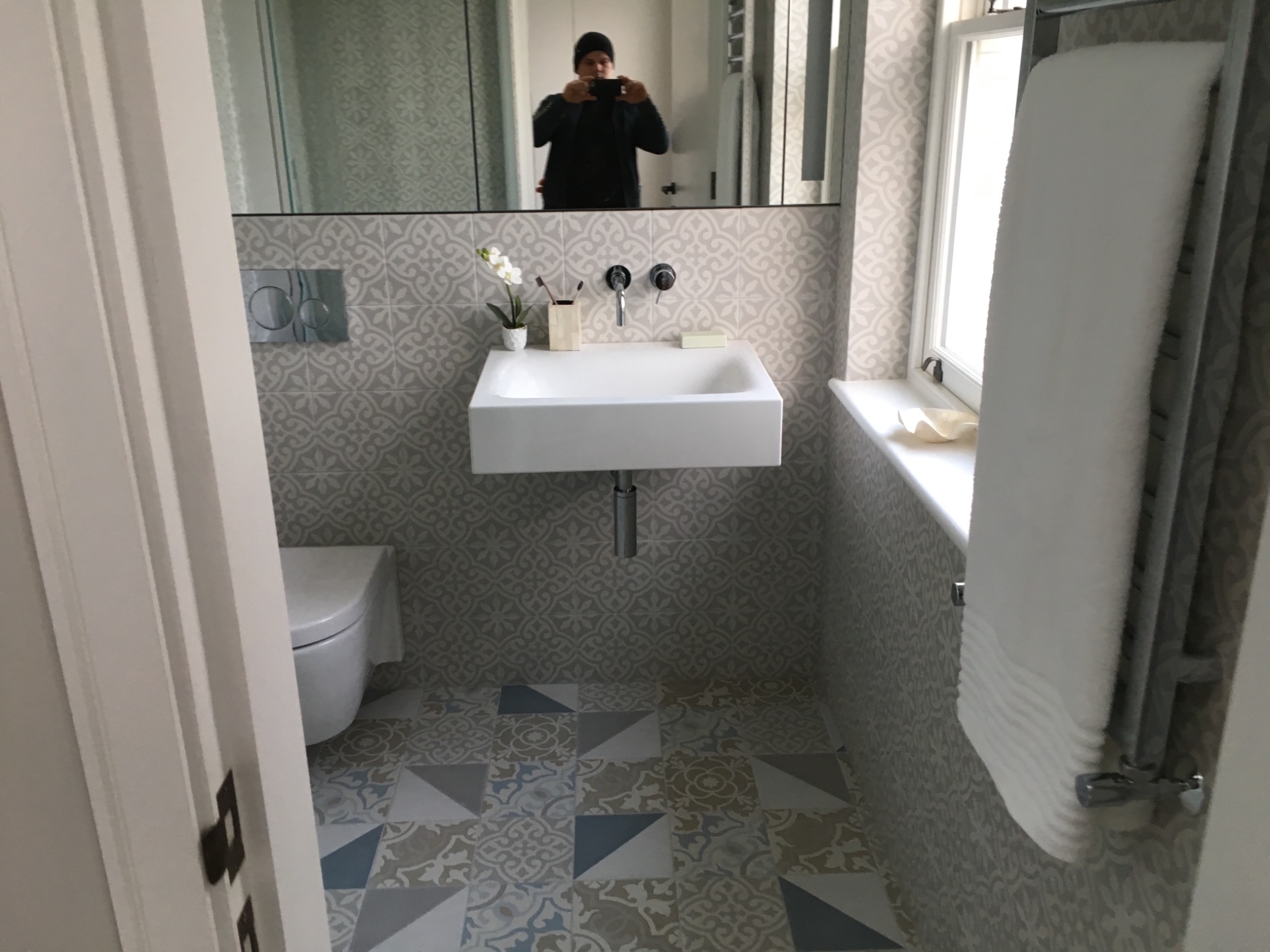 Nowoczesna łazienka z wiszącą umywalką, wzorzystymi płytkami na ścianach i podłodze, oraz grzejnikiem drabinkowym z białym ręcznikiem. W lustrze odbija się osoba robiąca zdjęcie.