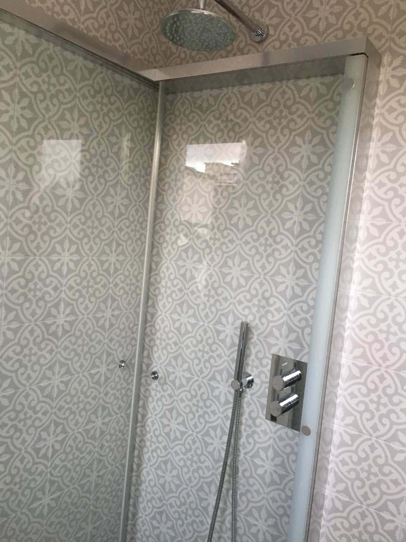 Szklana kabina prysznicowa z deszczownicą i baterią termostatyczną, wykończona płytkami w delikatny, szary wzór.