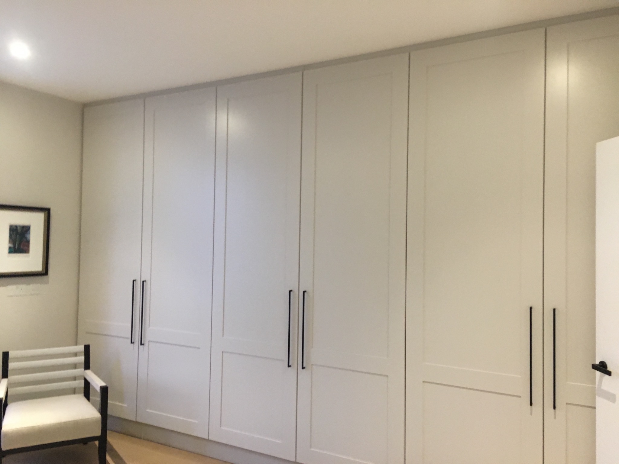 Wbudowana szafa z białymi, panelowymi drzwiami i długimi, czarnymi uchwytami, uzupełniona krzesłem z jasnym siedziskiem i czarną ramą oraz obrazem w czarnej ramie na ścianie.