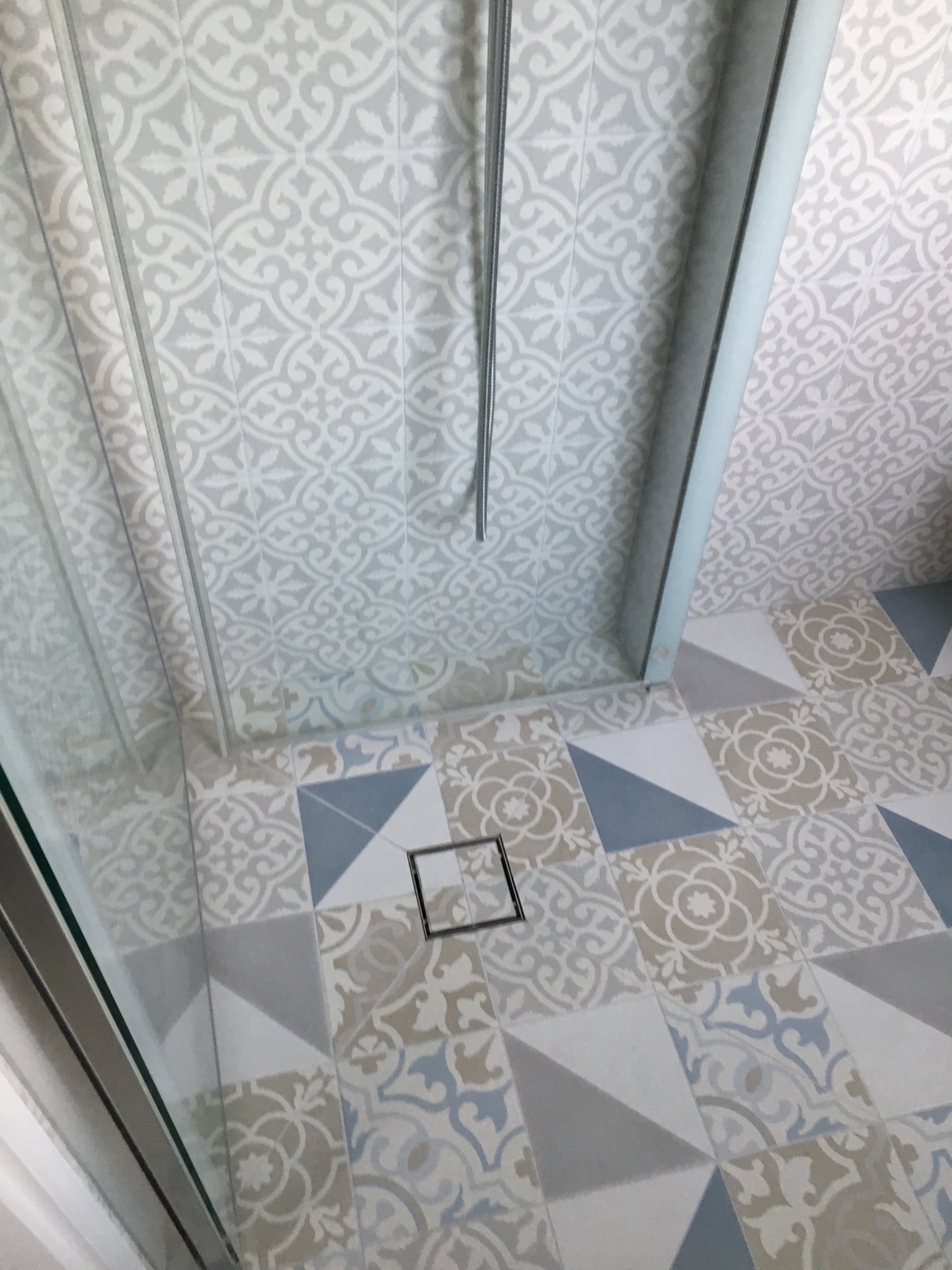 Nowoczesna kabina prysznicowa z odpływem liniowym w podłodze wyłożonej wzorzystymi płytkami w odcieniach szarości, błękitu i beżu.