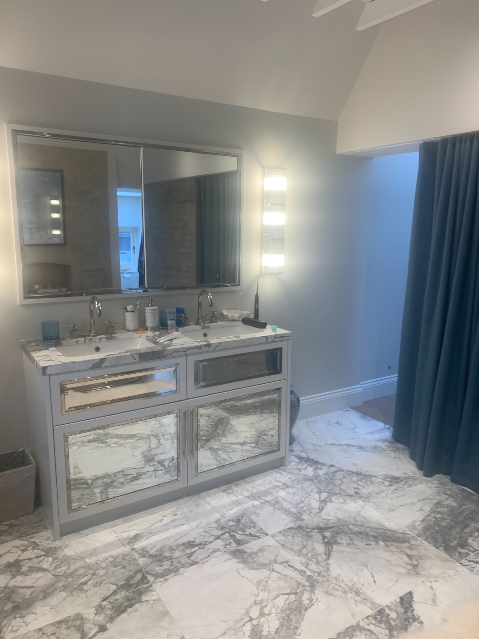 Elegancka łazienka z marmurową podłogą i szafką pod umywalką z lustrzanymi frontami, oświetlenie LED po bokach lustra, zasłona w tle.