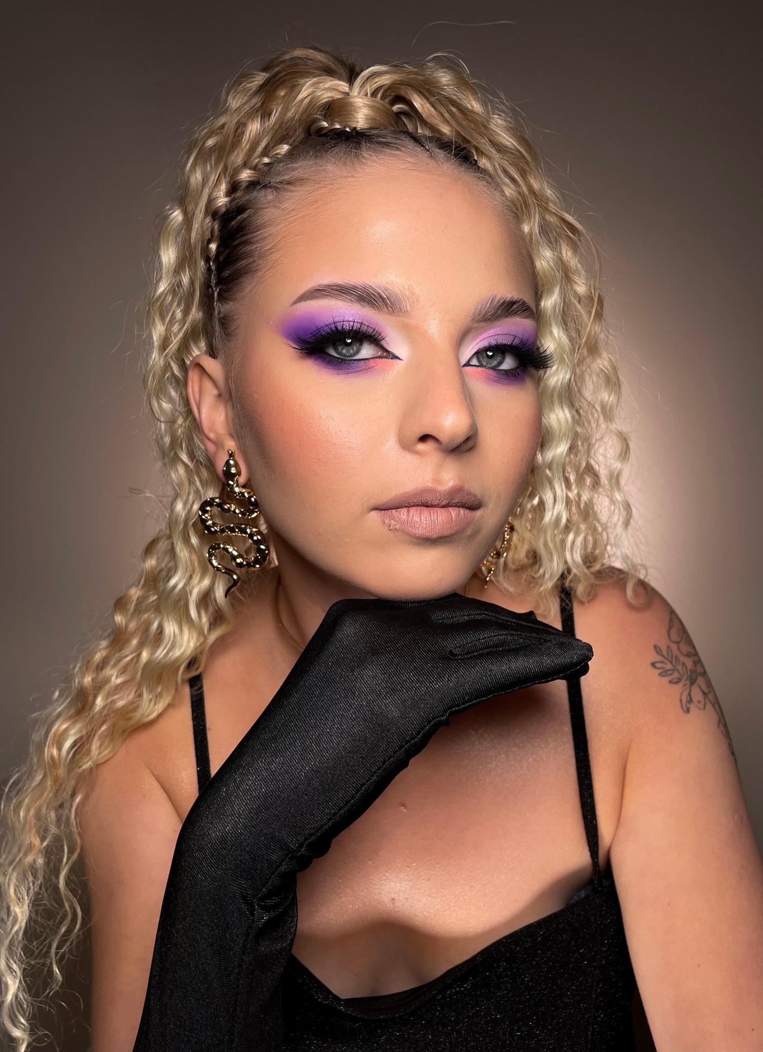 Portret kobiety z blond włosami upiętymi w warkocze, z intensywnym fioletowym cieniem na powiekach, długimi rzęsami, kolczykami w kształcie węży i czarną rękawiczką opartą o podbródek. Widoczny...