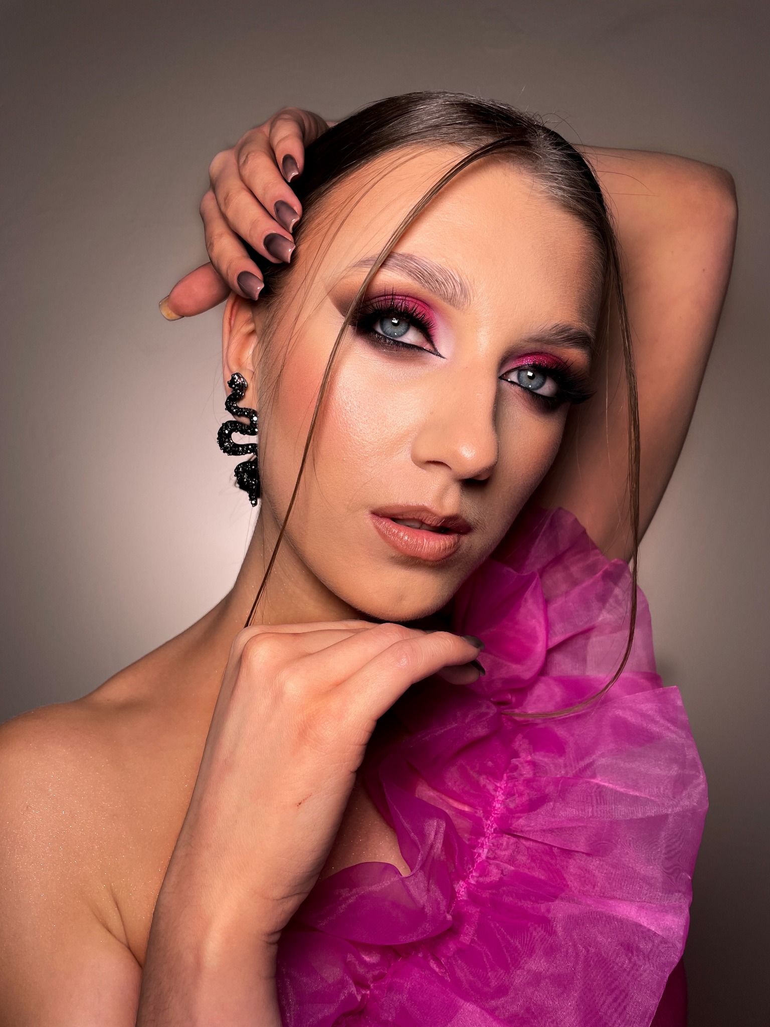 Portret kobiety z intensywnym różowym makijażem oczu, czarnymi kolczykami w kształcie węży i różową, marszczoną tkaniną, z rękami uniesionymi za głową, prezentujący efektowny wygląd.
