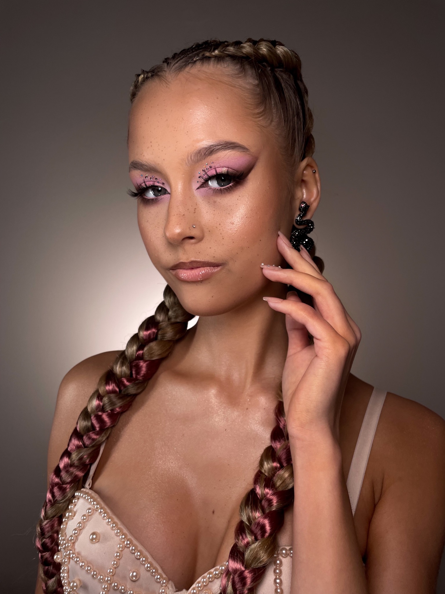 Portret kobiety z różowym makijażem oczu zdobionym brokatem, warkoczami z różowymi pasemkami i kolczykami w kształcie węża, dotykającej policzka.