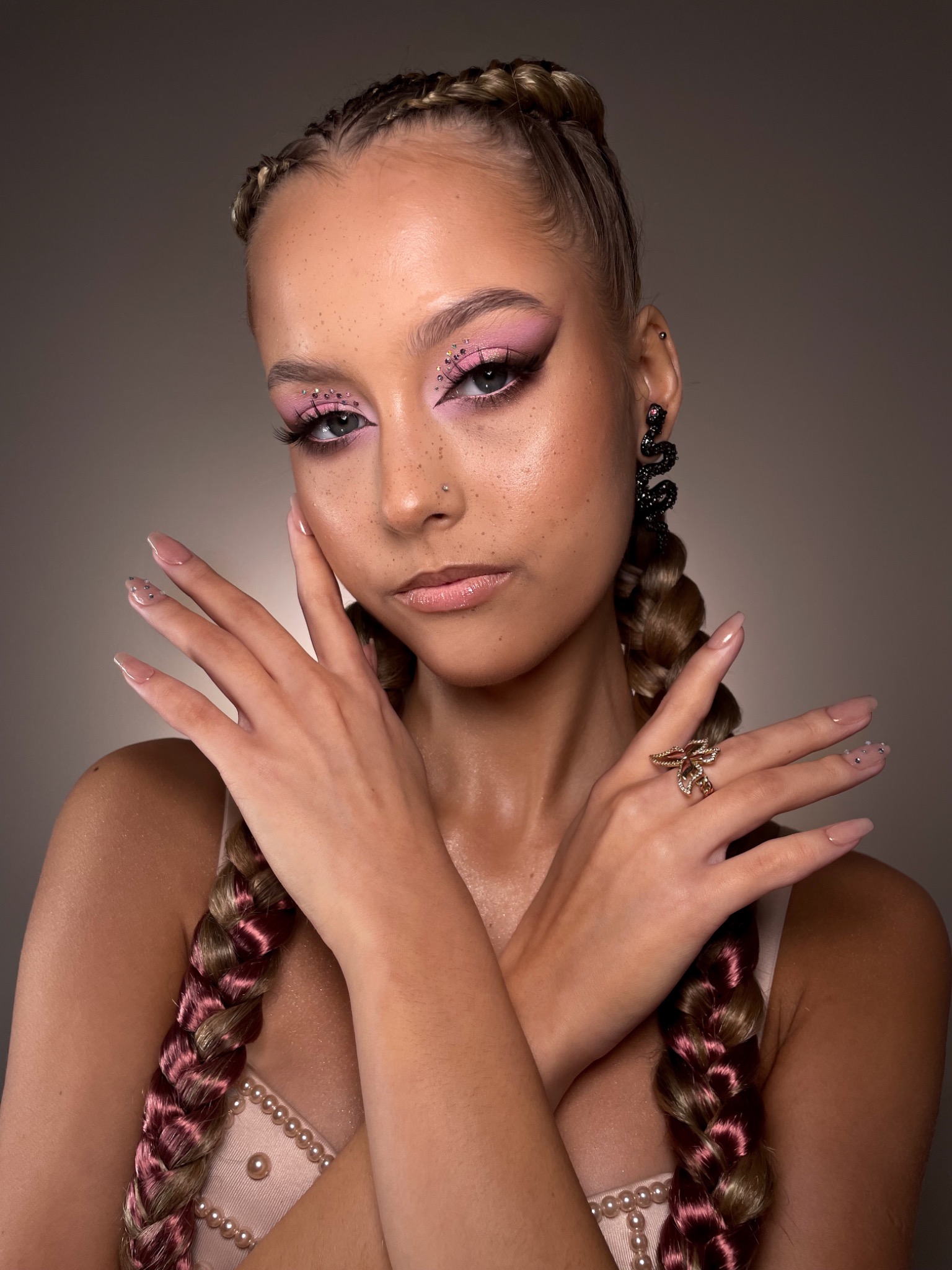Portret kobiety z różowym makijażem oczu, brokatem, warkoczami z różowymi wstawkami, ozdobnym pierścionkiem i kolczykami w kształcie węży.
