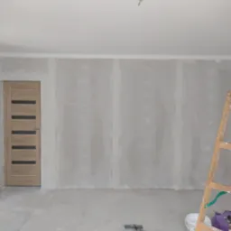 Pokój w trakcie remontu: ściany wyłożone płytami kartonowo-gipsowymi, drewniane drzwi z przeszkleniami, drabina oparta o ścianę, widoczne narzędzia i akcesoria malarskie.