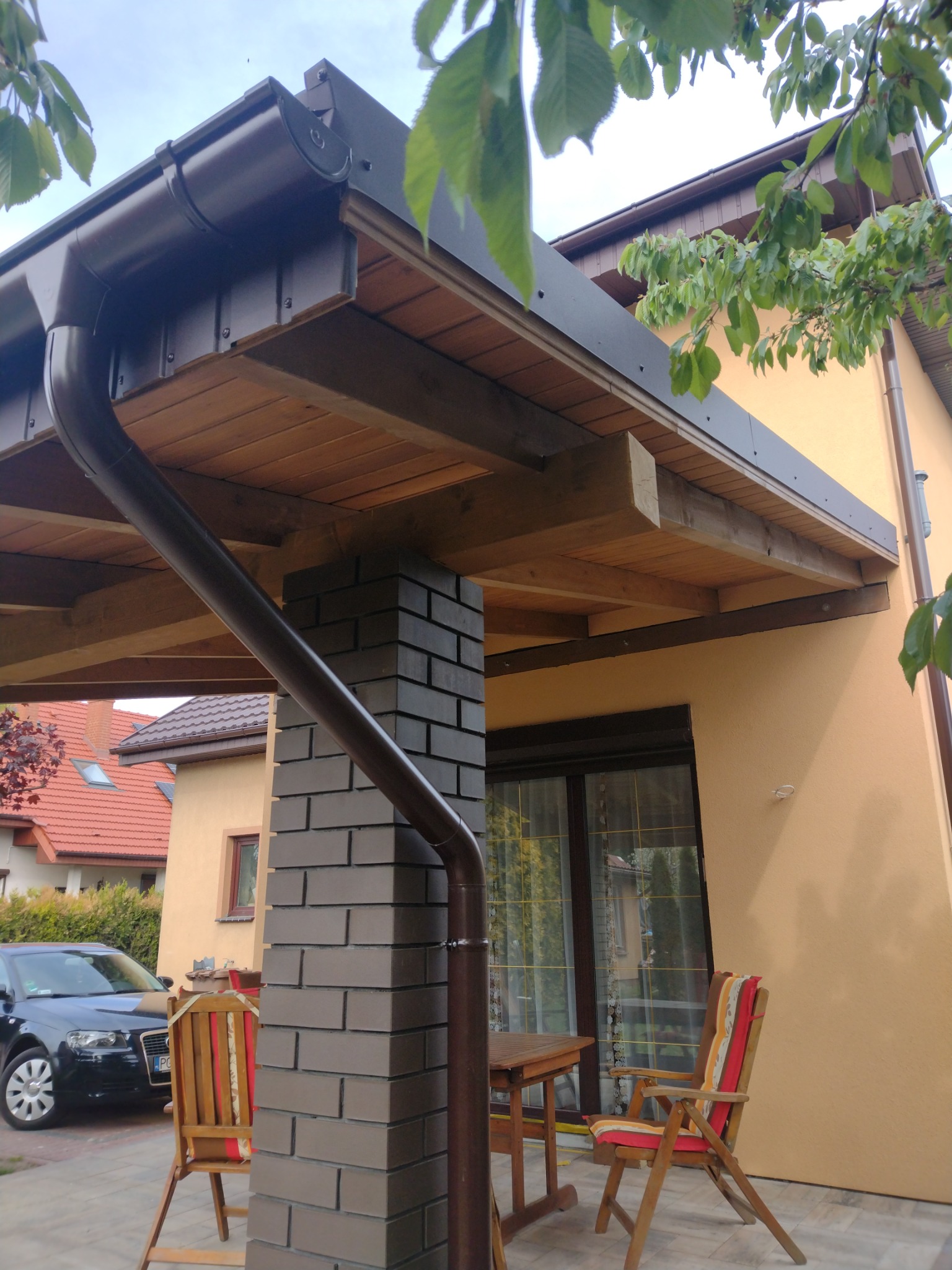 Zadaszenie tarasu z drewnianą konstrukcją, ceglanym filarem i brązowym systemem rynnowym, widoczne dwa krzesła z poduszkami i fragment samochodu.