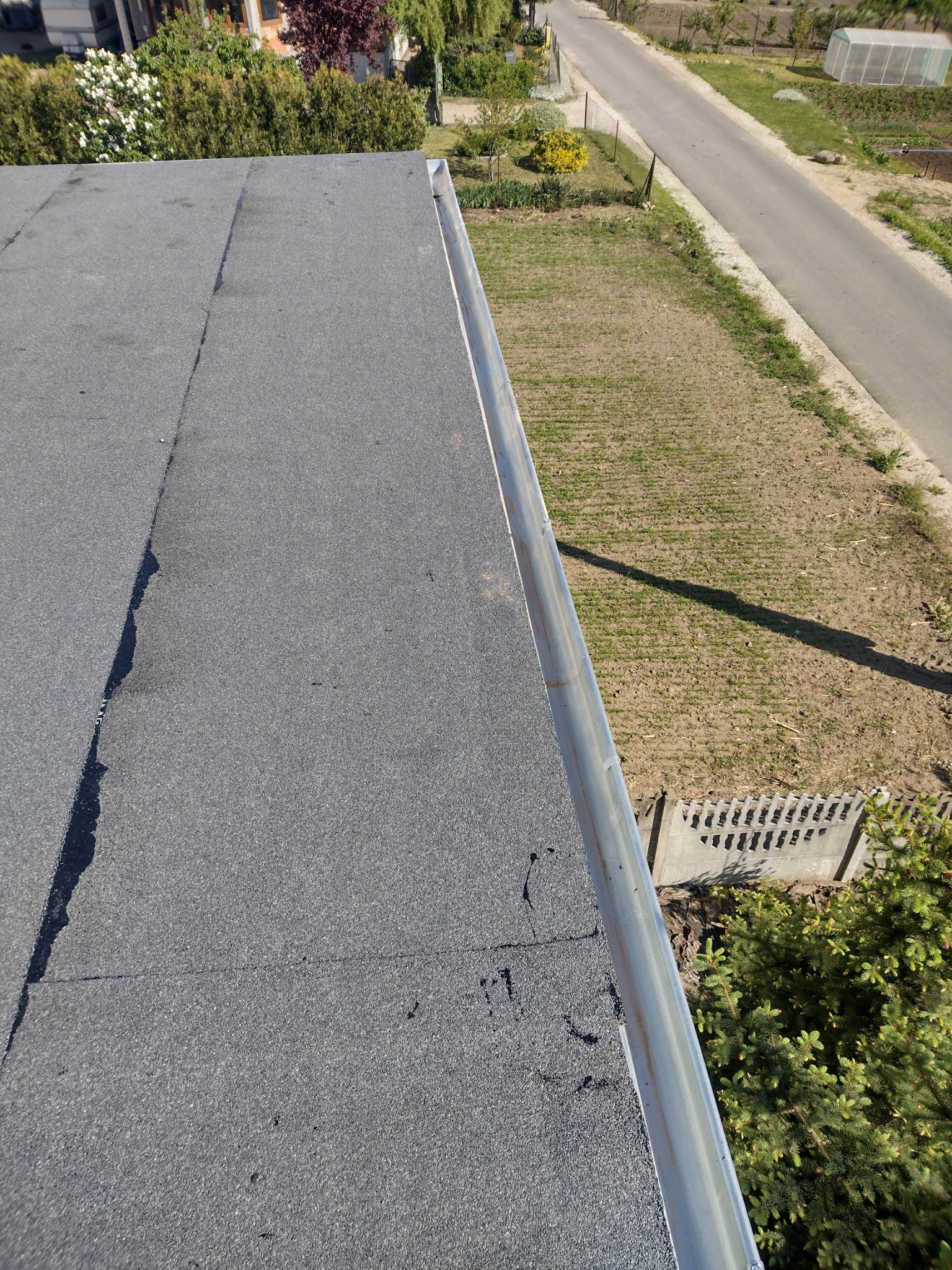 Widok z góry na płaski dach pokryty papą, z widoczną rynną i fragmentem ogrodu oraz drogi w tle.