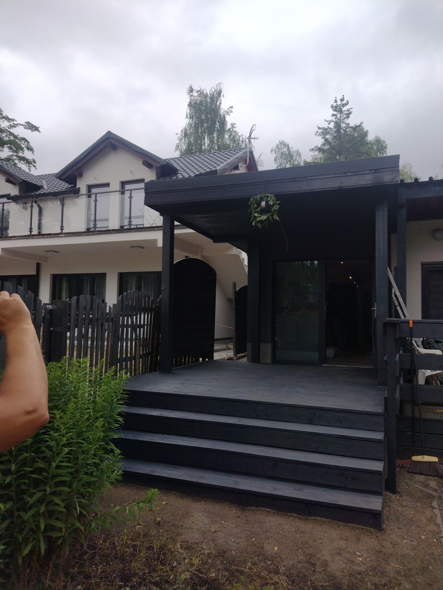 Nowoczesny taras z ciemnego drewna z zadaszeniem, widok na dwupiętrowy dom z balkonem, częściowo zasłonięty przez drewniany płot. W tle pochmurne niebo i drzewa.