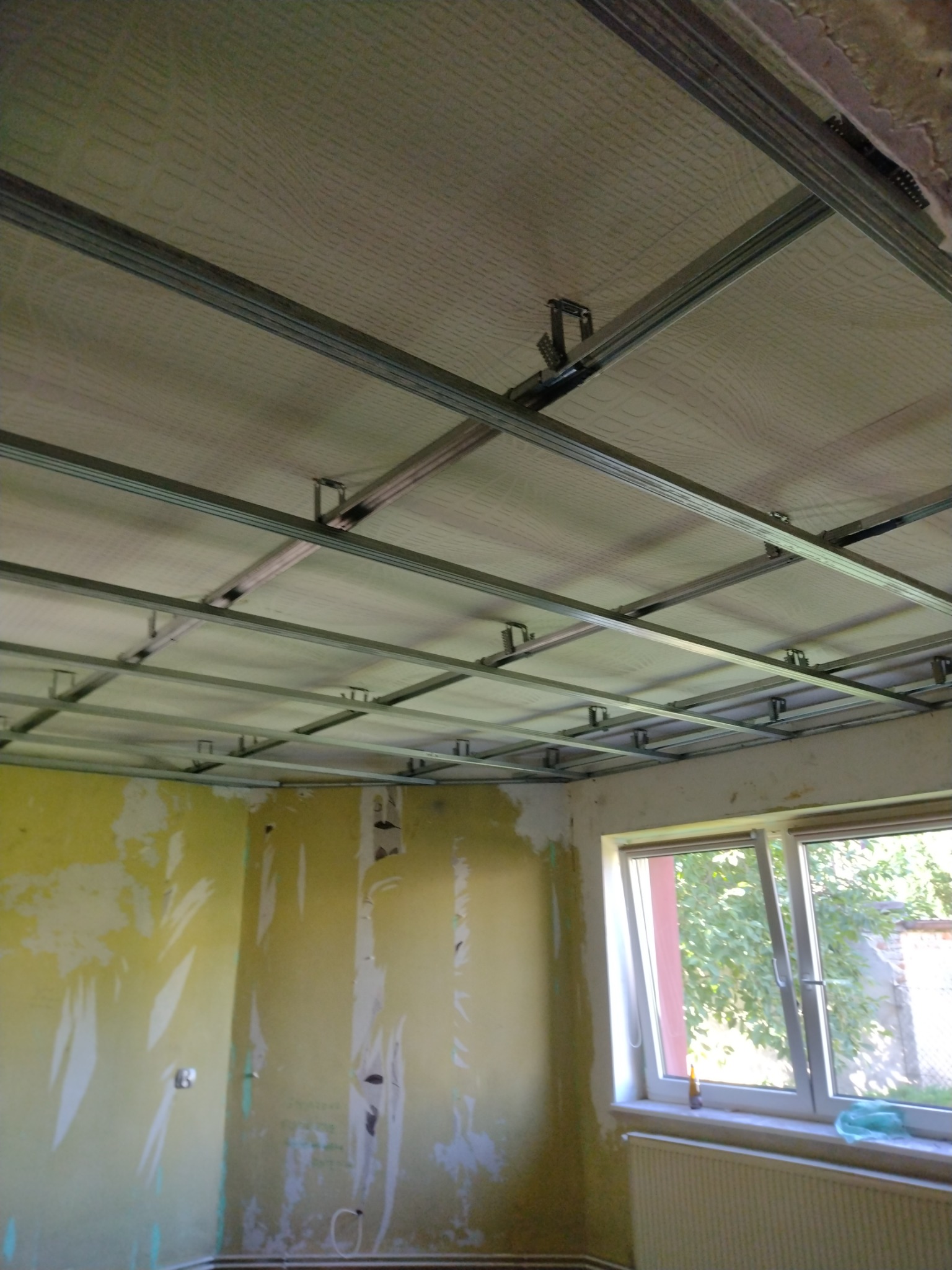 Montaż stelaża sufitu podwieszanego w pomieszczeniu z przygotowanymi ścianami, widoczne profile metalowe i płyty.