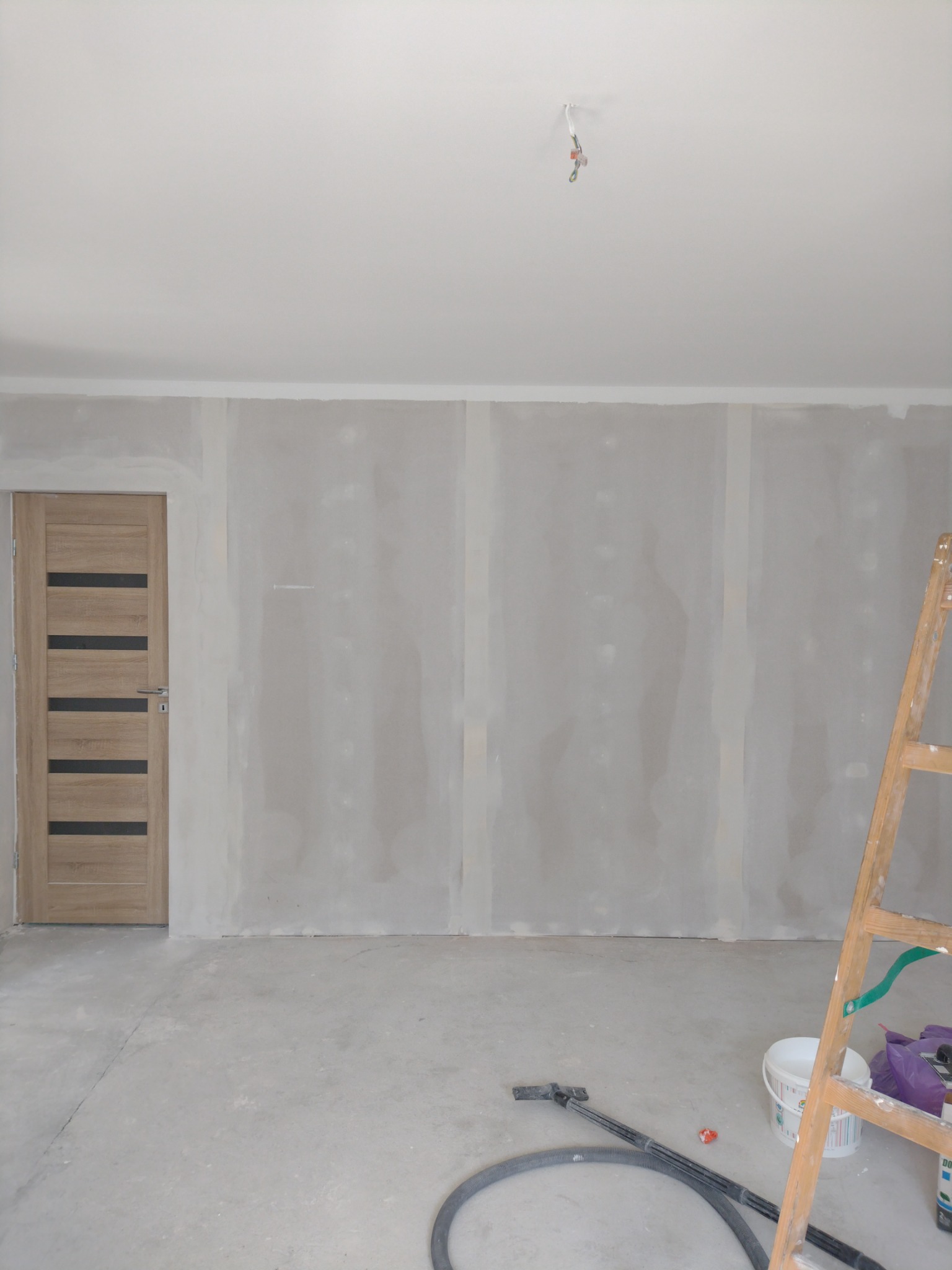 Pokój w trakcie remontu: ściany wyłożone płytami kartonowo-gipsowymi, drewniane drzwi z przeszkleniami, drabina oparta o ścianę, widoczne narzędzia i akcesoria malarskie.