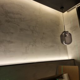 WEGA - Ściana z dekoracyjną, szarą fakturą, oświetlona listwą LED, z wiszącą lampą w kształcie szarej kuli i zielonymi liniami lasera krzyżowego.