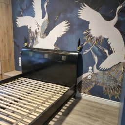 WEGA - Ściana z motywem żurawi za czarną ramą łóżka z drewnianym stelażem, widoczna puszka z farbą na podłodze.