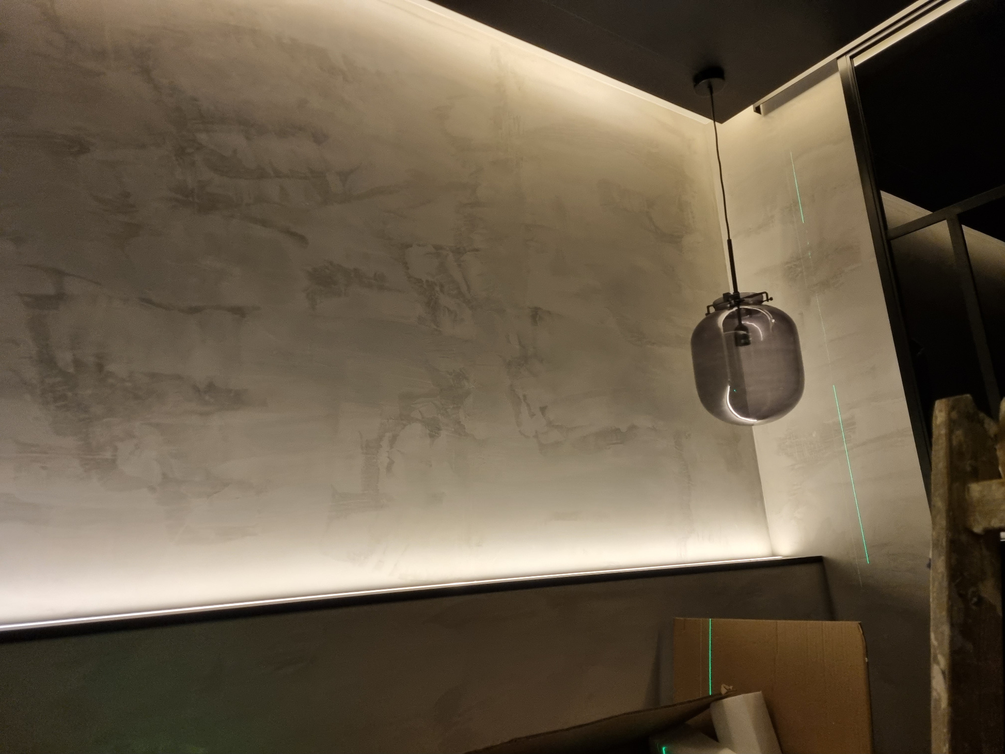 Ściana z dekoracyjną, szarą fakturą, oświetlona listwą LED, z wiszącą lampą w kształcie szarej kuli i zielonymi liniami lasera krzyżowego.