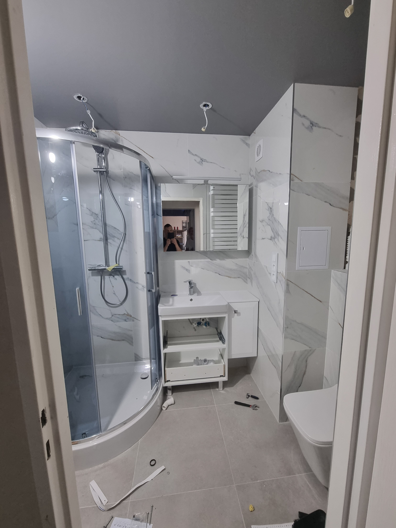 Niedokończona łazienka z kabiną prysznicową, umywalką z szafką i podwieszaną toaletą. Widoczne narzędzia i niedociągnięcia wykończeniowe, szare płytki podłogowe i białe płytki imitujące marmur...