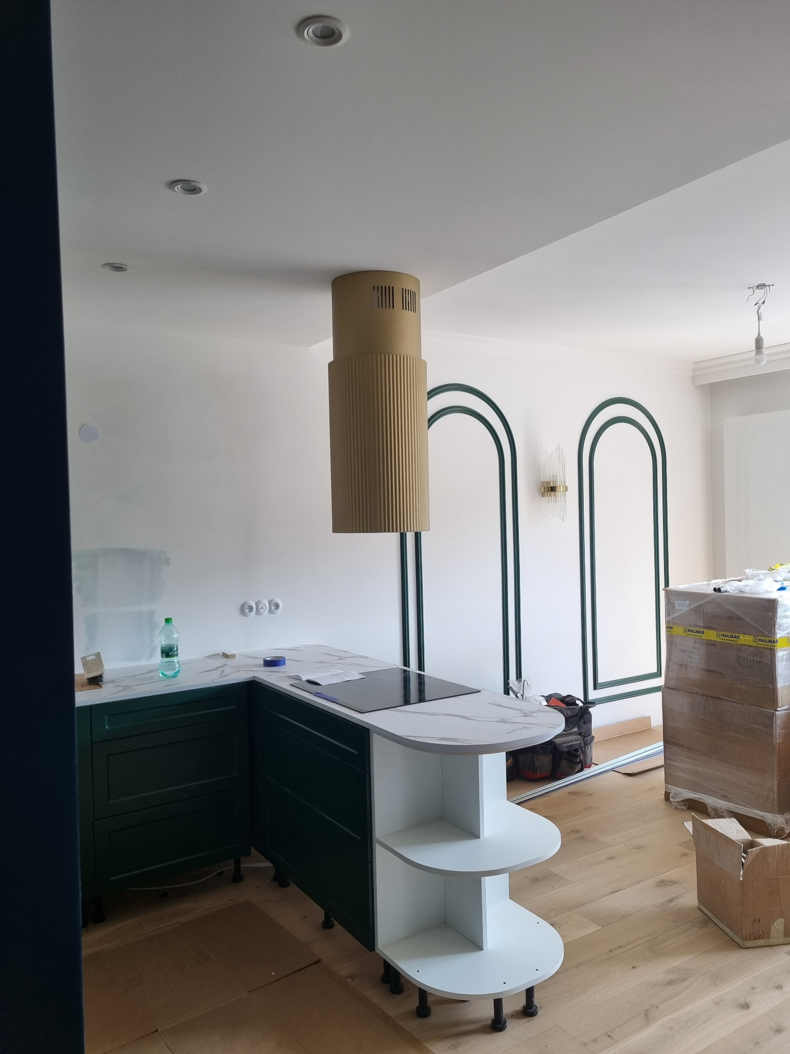 Nowoczesna kuchnia w trakcie montażu: ciemnozielone szafki, marmurowy blat, okrągły półwysep z białymi półkami i złoty okap z żłobieniami. Na ścianie białe listwy w kształcie łuków.